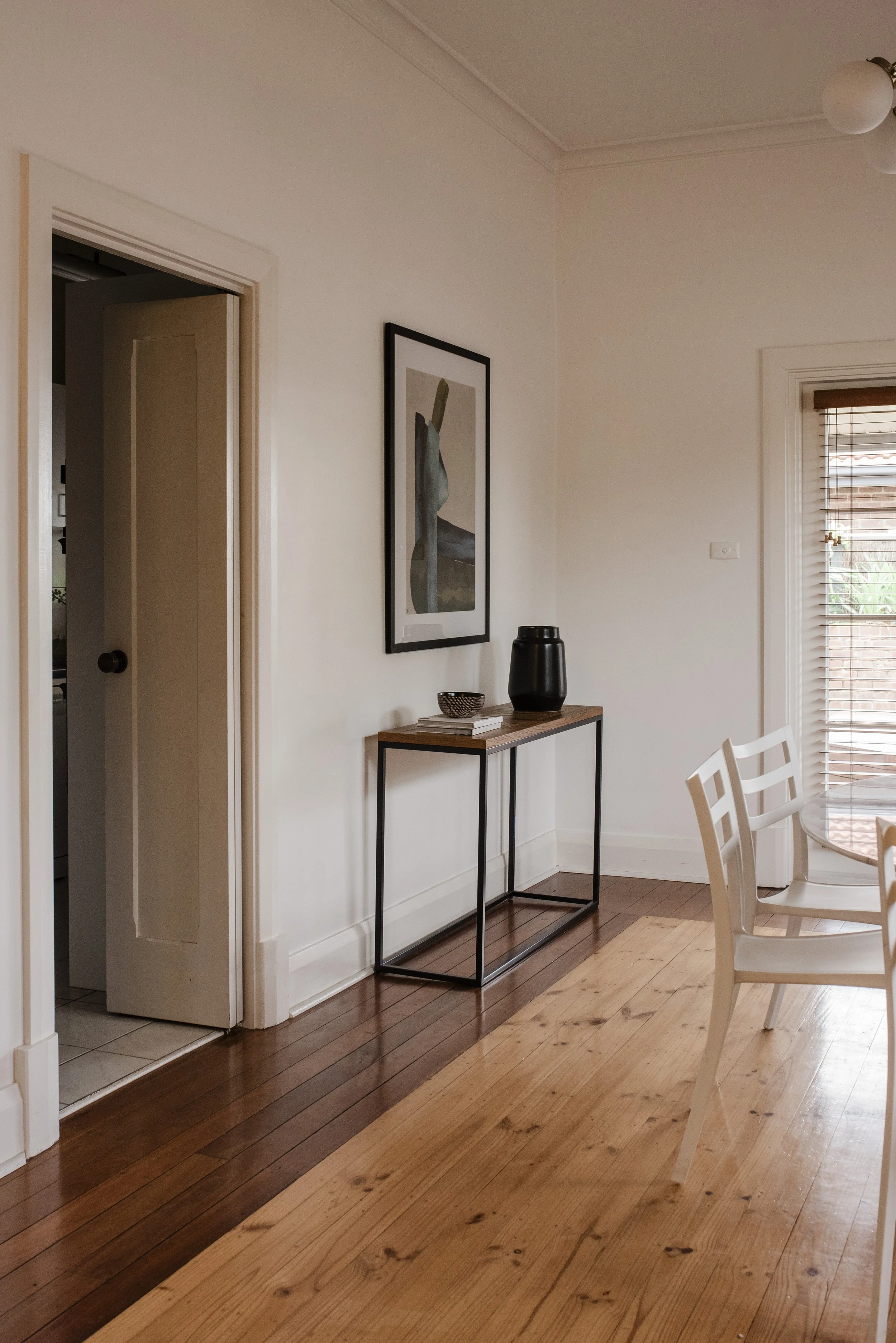 Interior of a room with wooden flooring, a narrow black metal-framed console table with a wooden top, placed against a white wall. On the table are a black vase, a small bowl, and a few books. Above the table hangs a vertical rectangular framed artwo