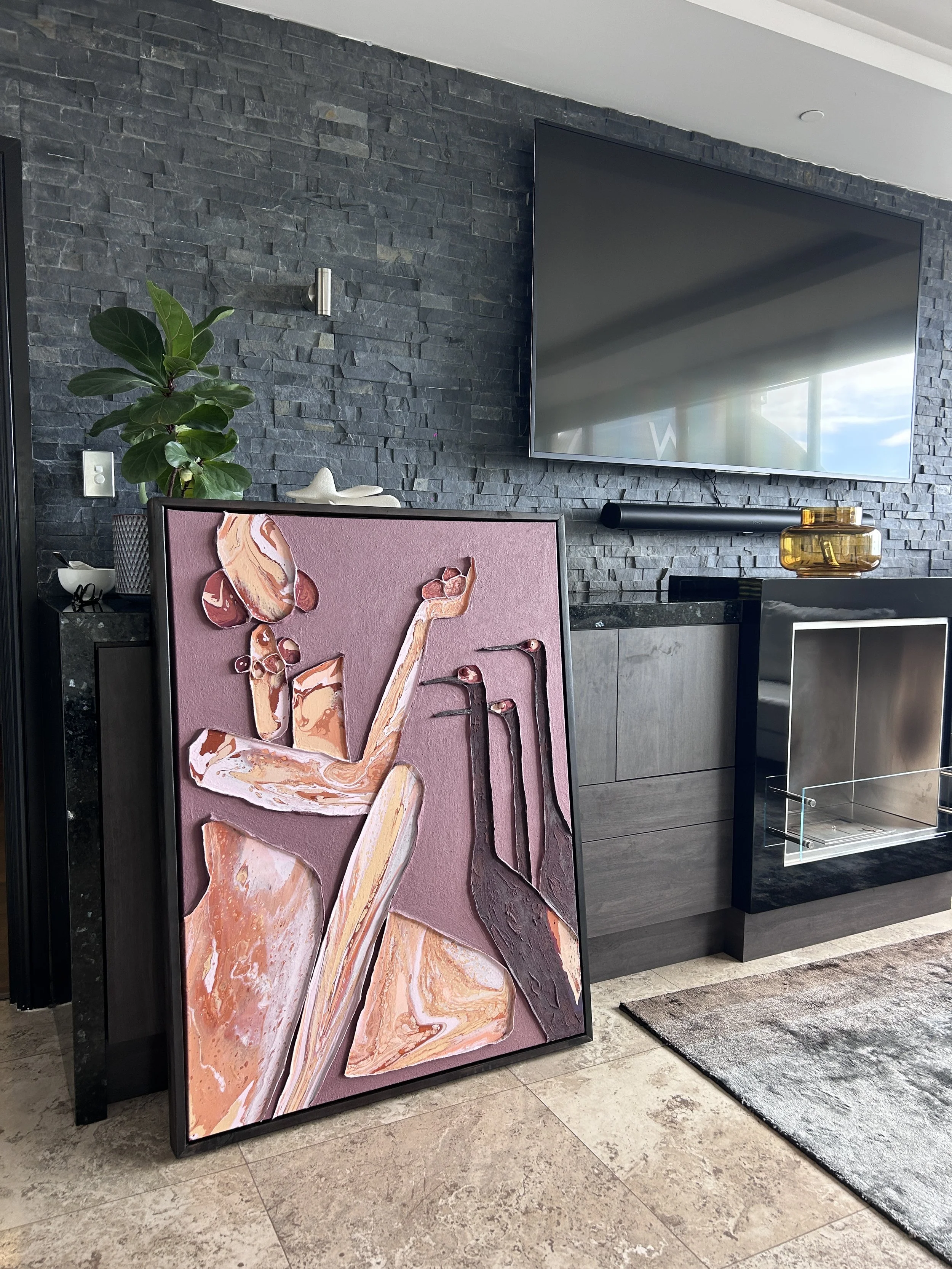 A modern living room corner with a large flat-screen TV mounted on a dark stone wall, a potted plant, a piece of artwork with a pink background and a stylized woman, a yellow glass decorative item, and a black cabinet surrounding the TV, with a light