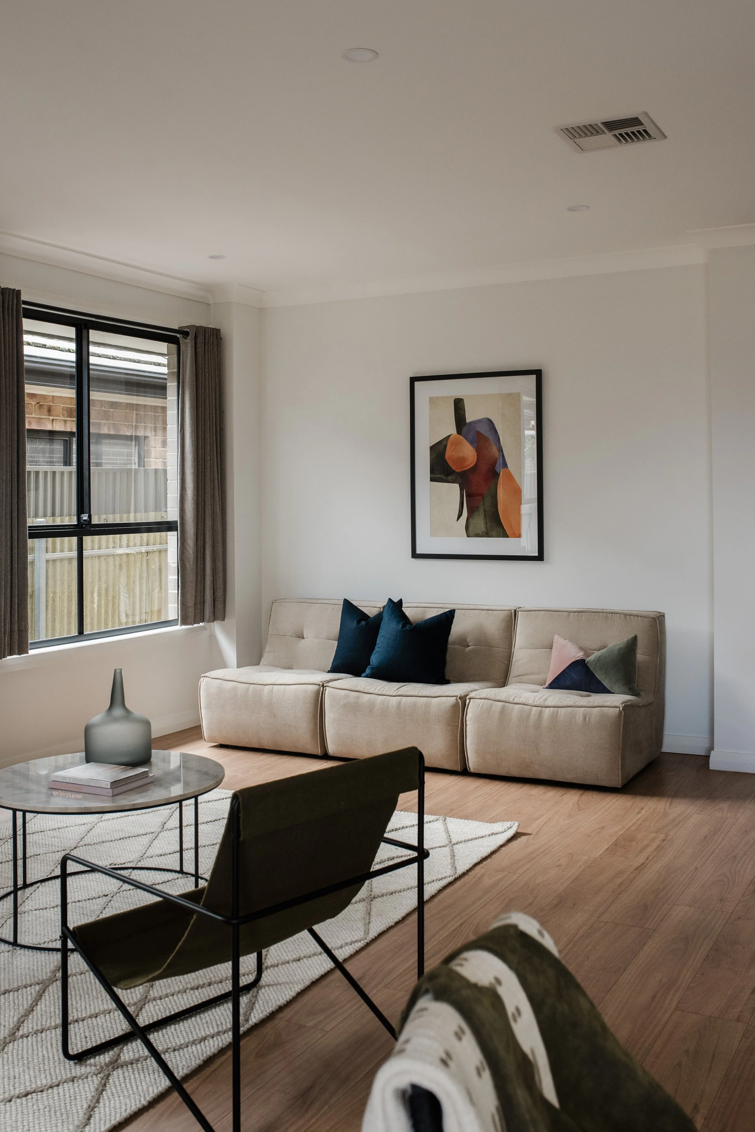 Living room with a cream sofa adorned with dark blue and pink cushions, a glass coffee table with a decorative vase and books, a textured area rug, a framed abstract art piece on the white wall, a window with beige curtains, and hardwood flooring. Pe