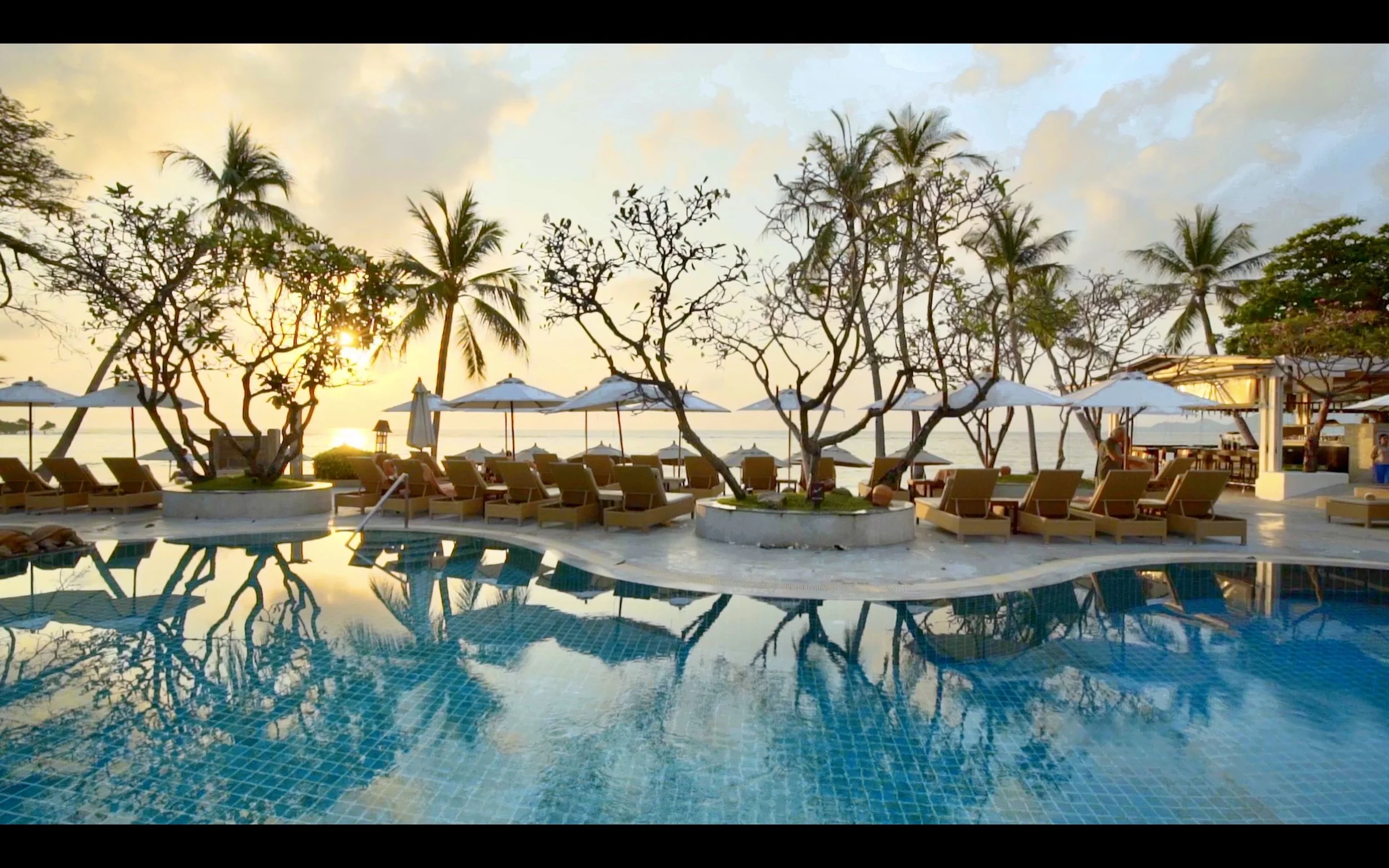 Swimming pool at sunset with lounge chairs, umbrellas, palm trees, and a bar area nearby on a tropical beach.