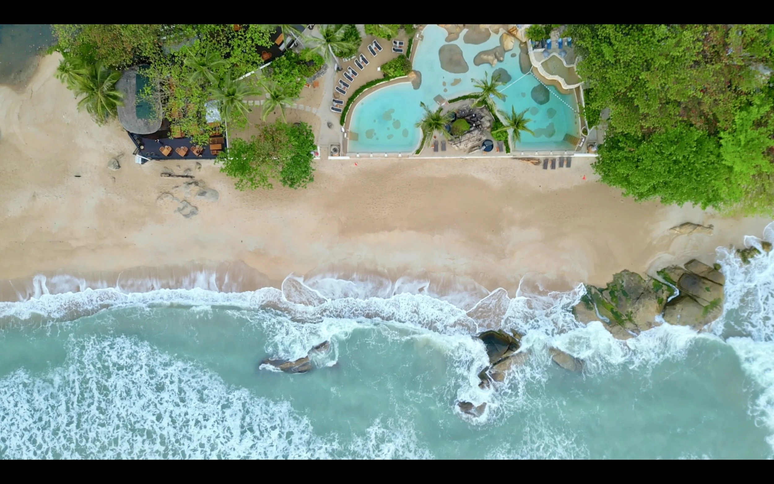 Aerial view of a beach with sand, ocean waves crashing against rocks, and a swimming pool area surrounded by trees and lounge chairs.