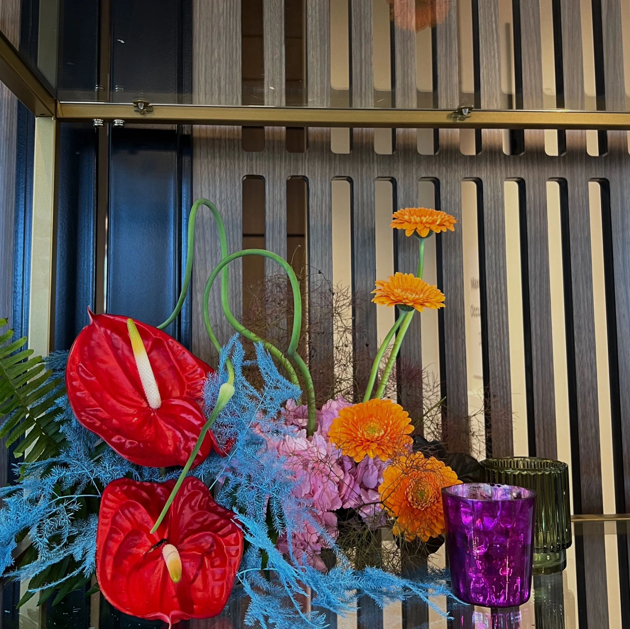 Colorful floral arrangement with red anthuriums, orange daisies, pink and purple flowers, blue foliage, and purple and green glass candle holders on a reflective surface with a wooden slatted background.