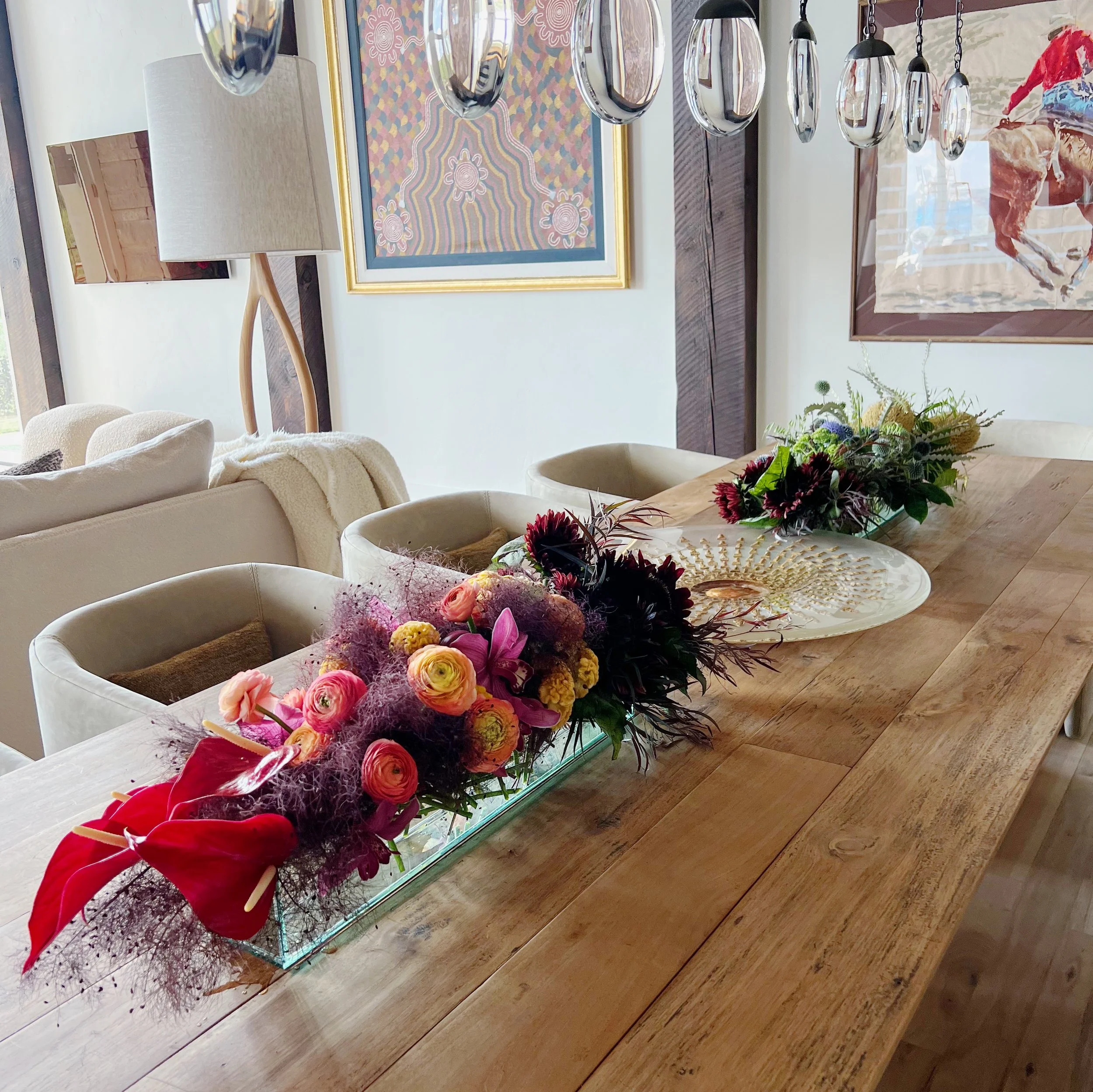 Dining table with colorful flower arrangements, wooden surface, modern hanging lights, and framed artwork on the wall.