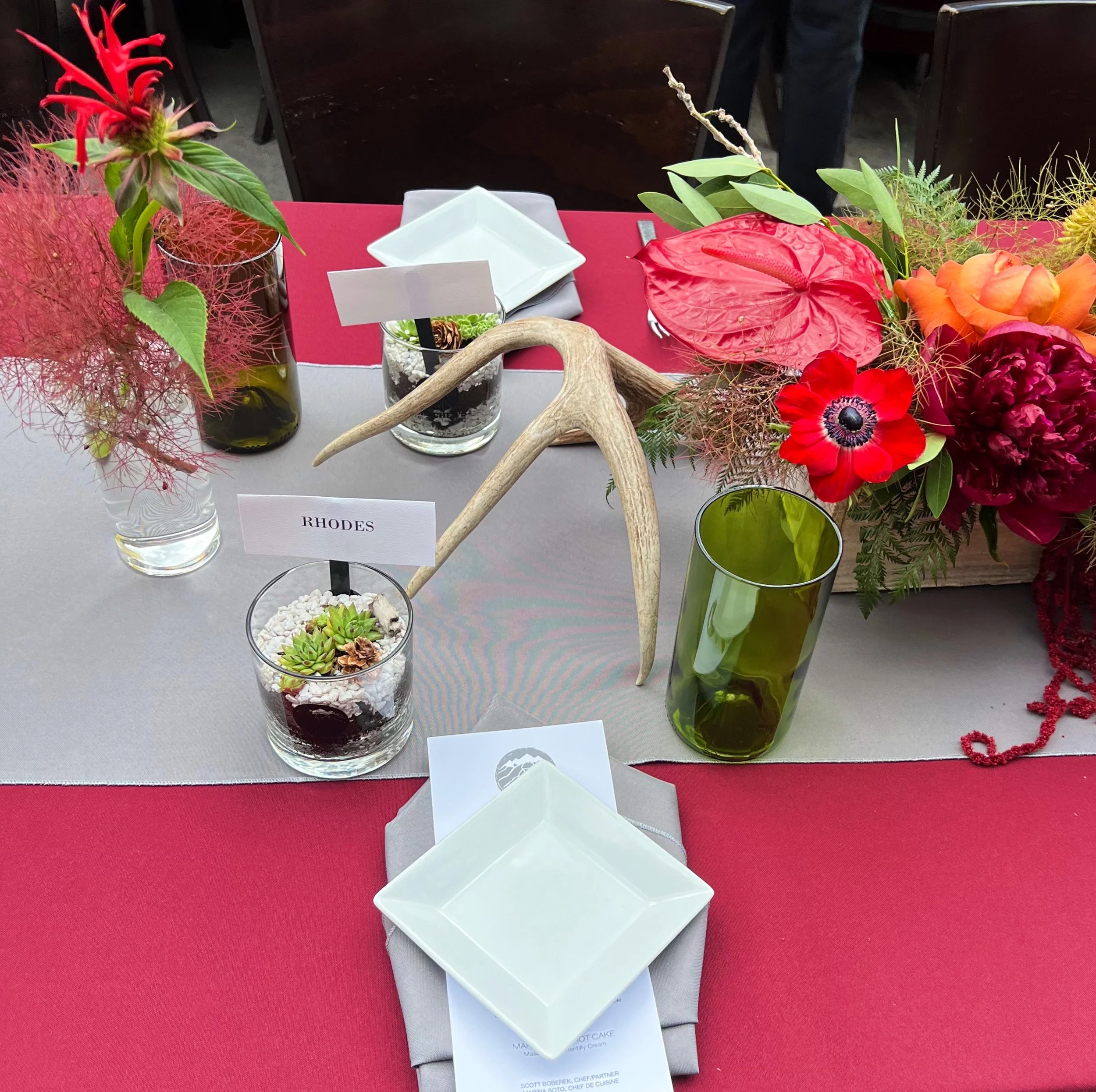 Table setting with red tablecloth, grey table runner, small square plates, green glass cups, and decorative elements including floral arrangements, succulents in glass containers, and antlers.