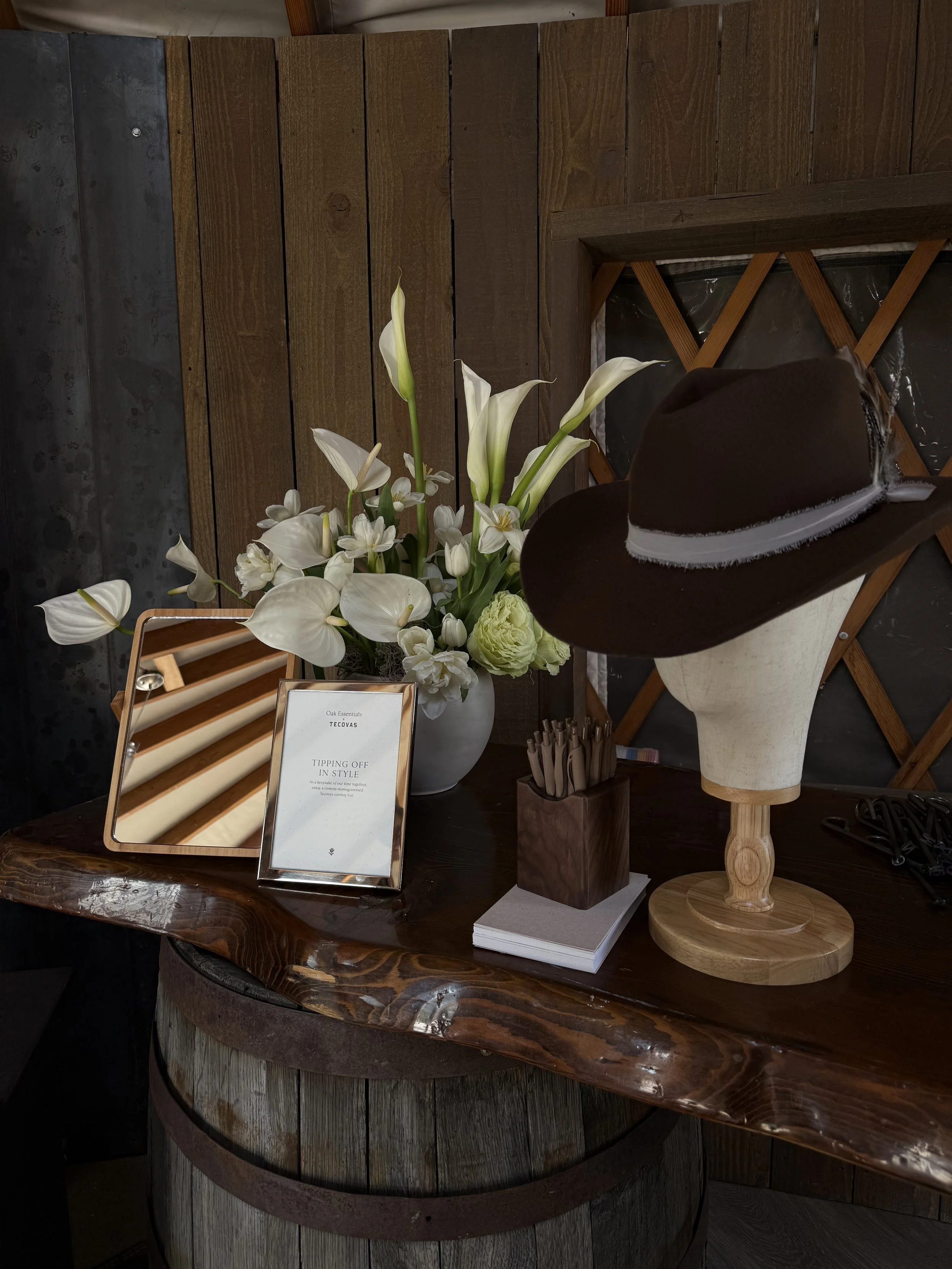 A wooden table with a flower arrangement of white calla lilies and other white flowers, a mirror, a small framed sign, a stack of white paper, a wooden container holding wooden stir sticks, and a mannequin head wearing a brown hat with a white feathe