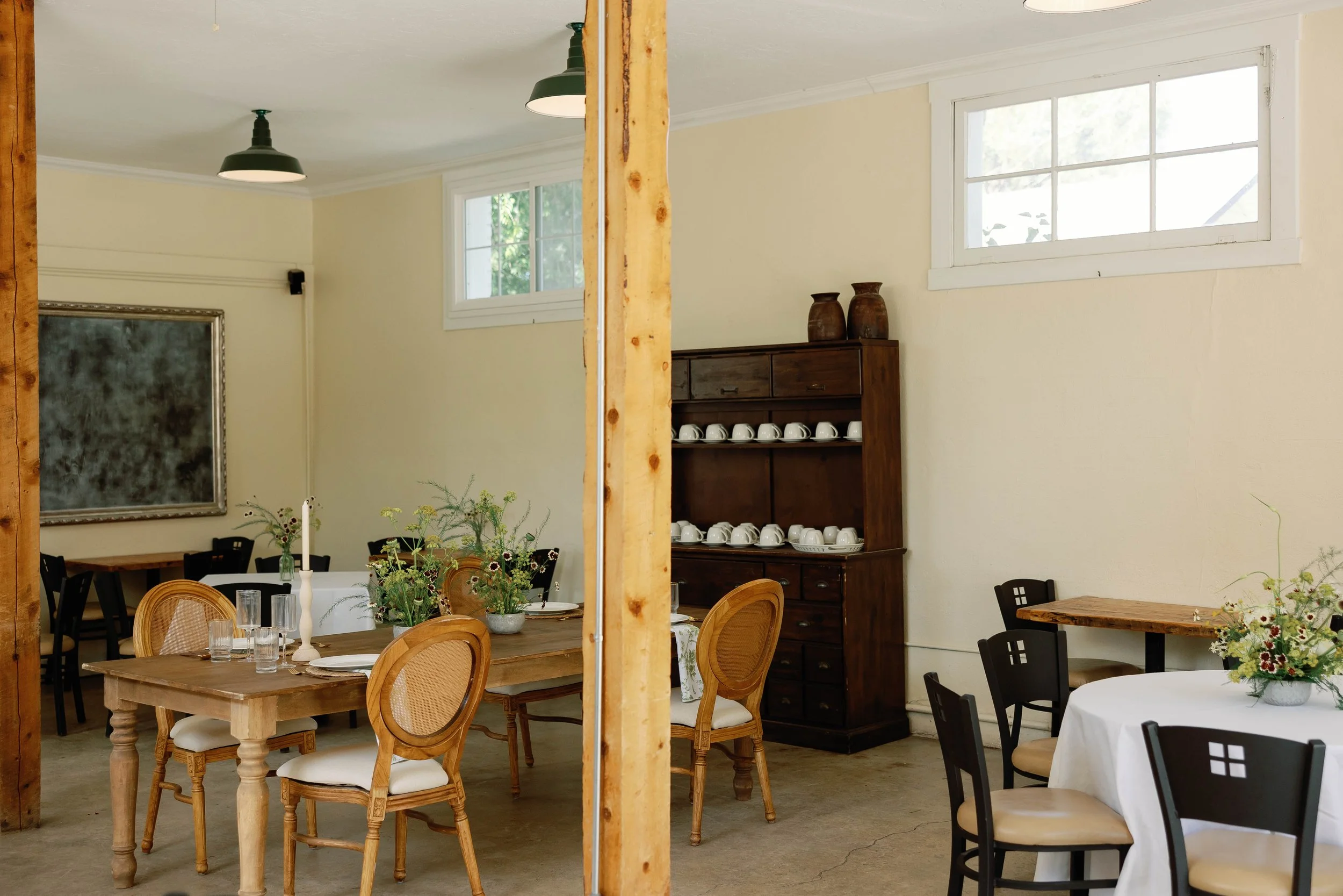 A dining room with wooden tables and chairs, decorated with floral centerpieces, displaying an empty space divided by a wooden beam, with a vintage cabinet holding white cups and two vases, and windows letting in natural light.