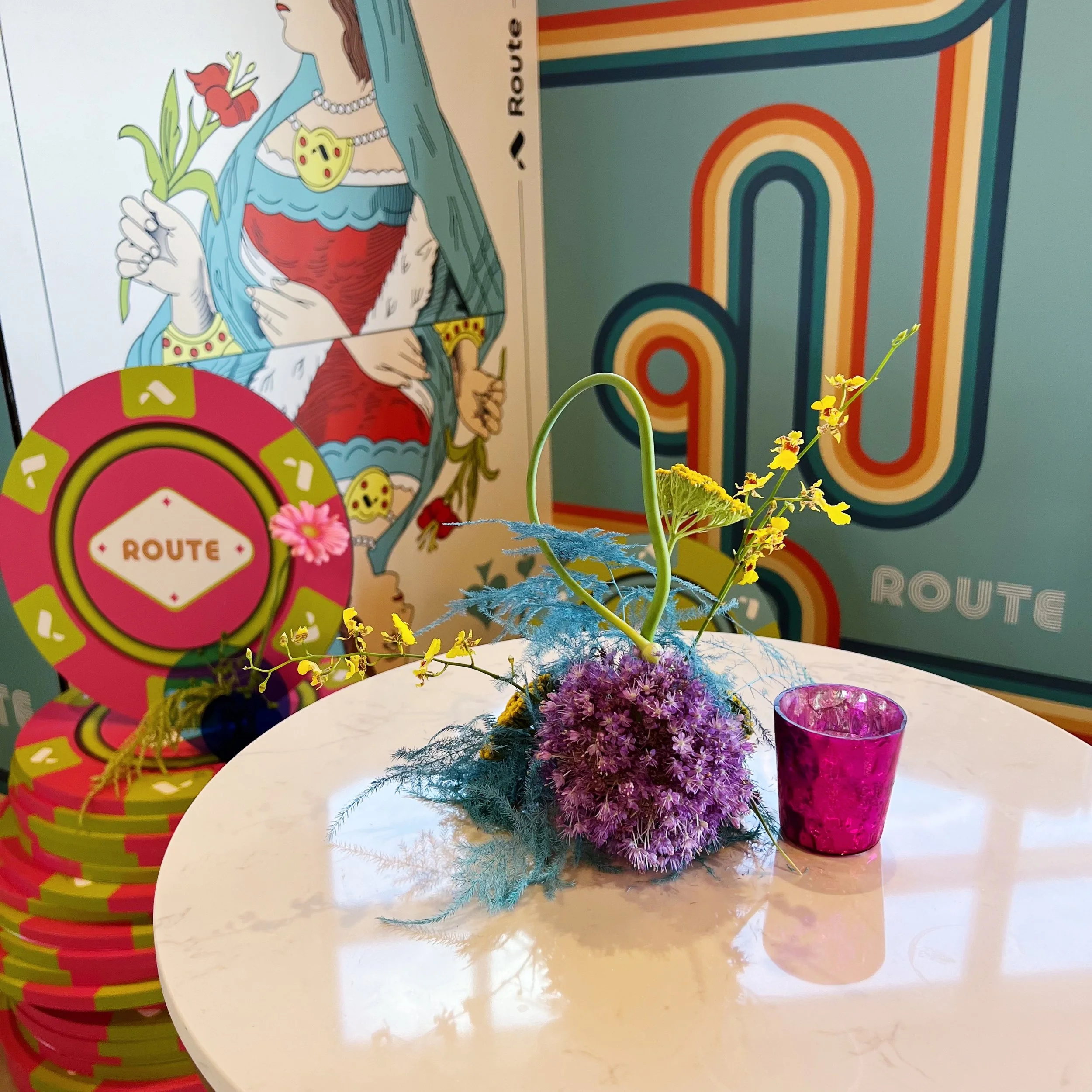 A floral arrangement with purple and yellow flowers and green foliage in a blue vase, placed on a white table next to a pink candle holder. Behind the table, there are colorful poker chips with the word 'ROUTE' and a large illustrated poster of a wom