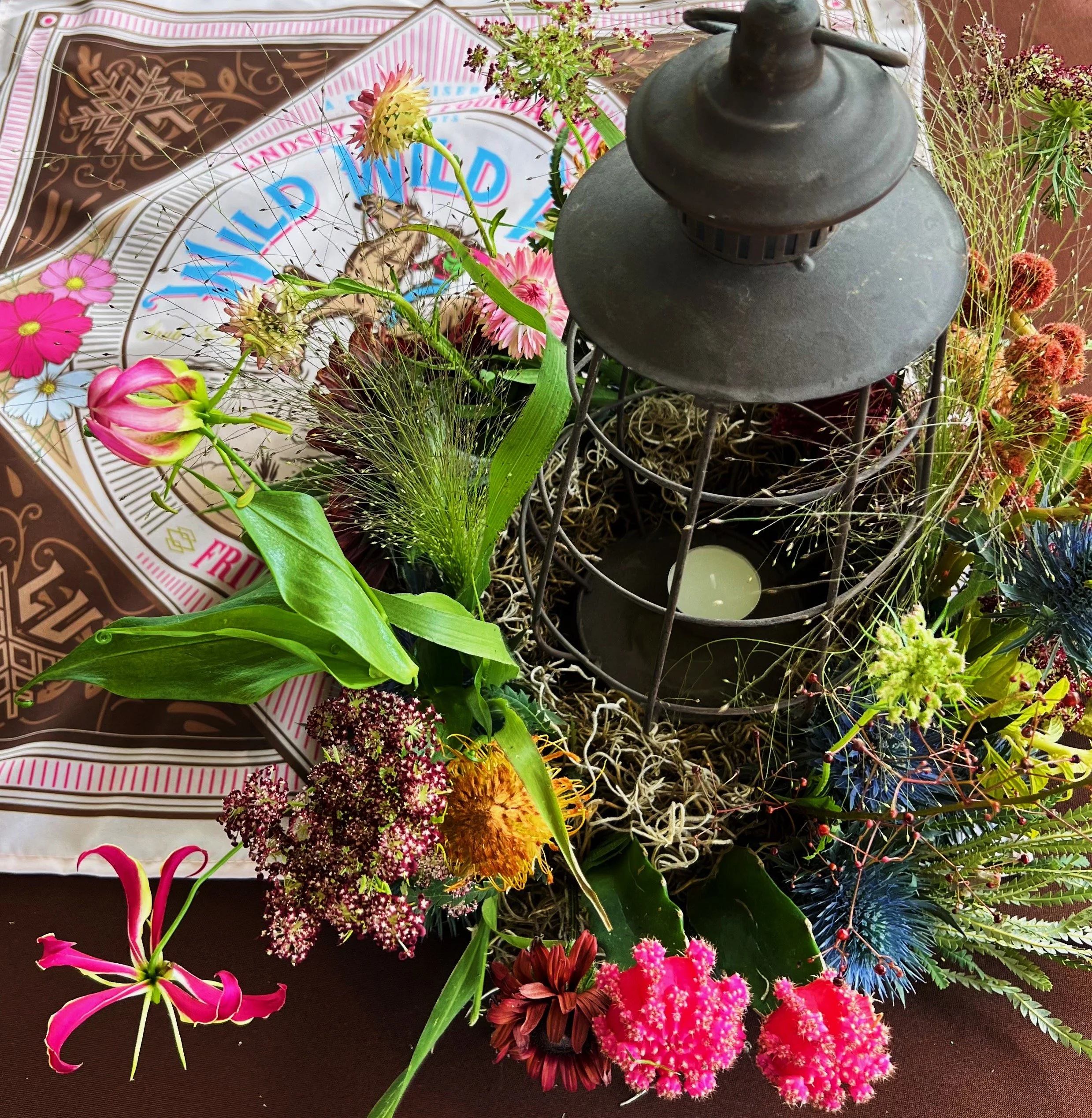 A decorative arrangement with a black metal lantern containing a white candle, surrounded by an assortment of vibrant flowers and greenery. A colorful printed textile with floral designs is partly visible in the background.