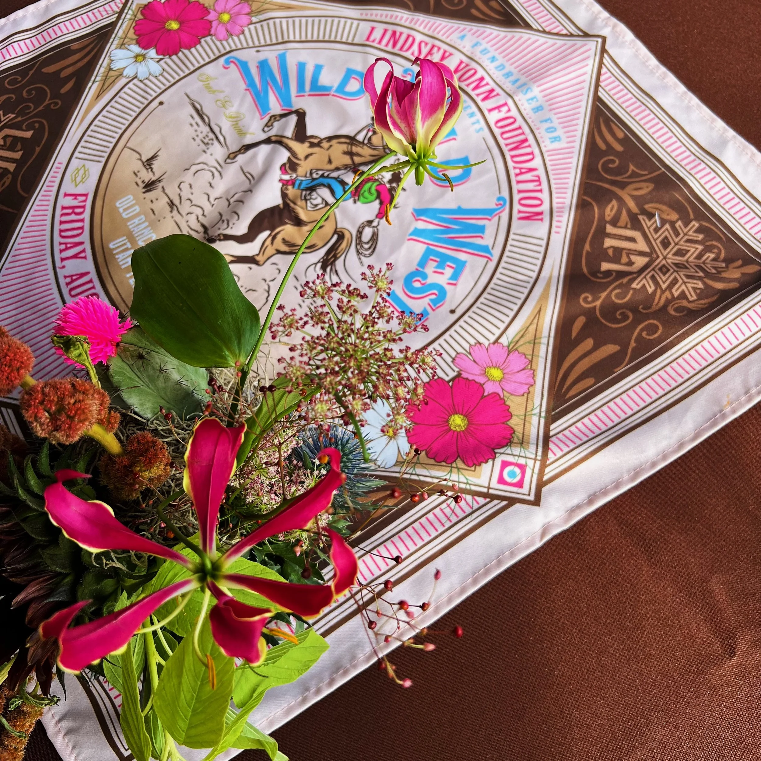 Decorative scarf with "Wild Wild West" text, featuring floral designs and a central image of a horse, paired with a vibrant bouquet of pink flowers and greenery.