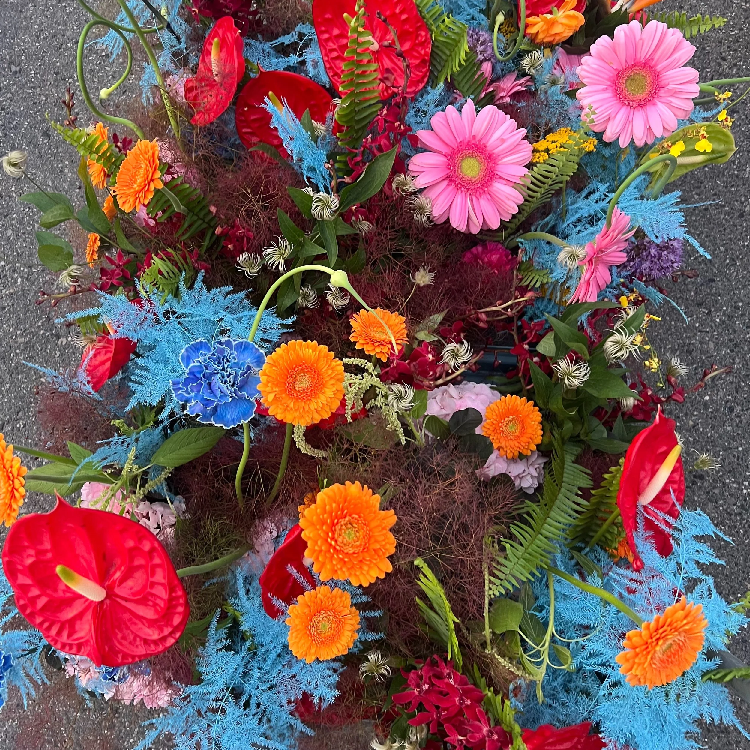 Colorful floral arrangement with pink gerbera daisies, orange marigolds, red anthuriums, blue delphiniums, and green foliage on a dark surface.