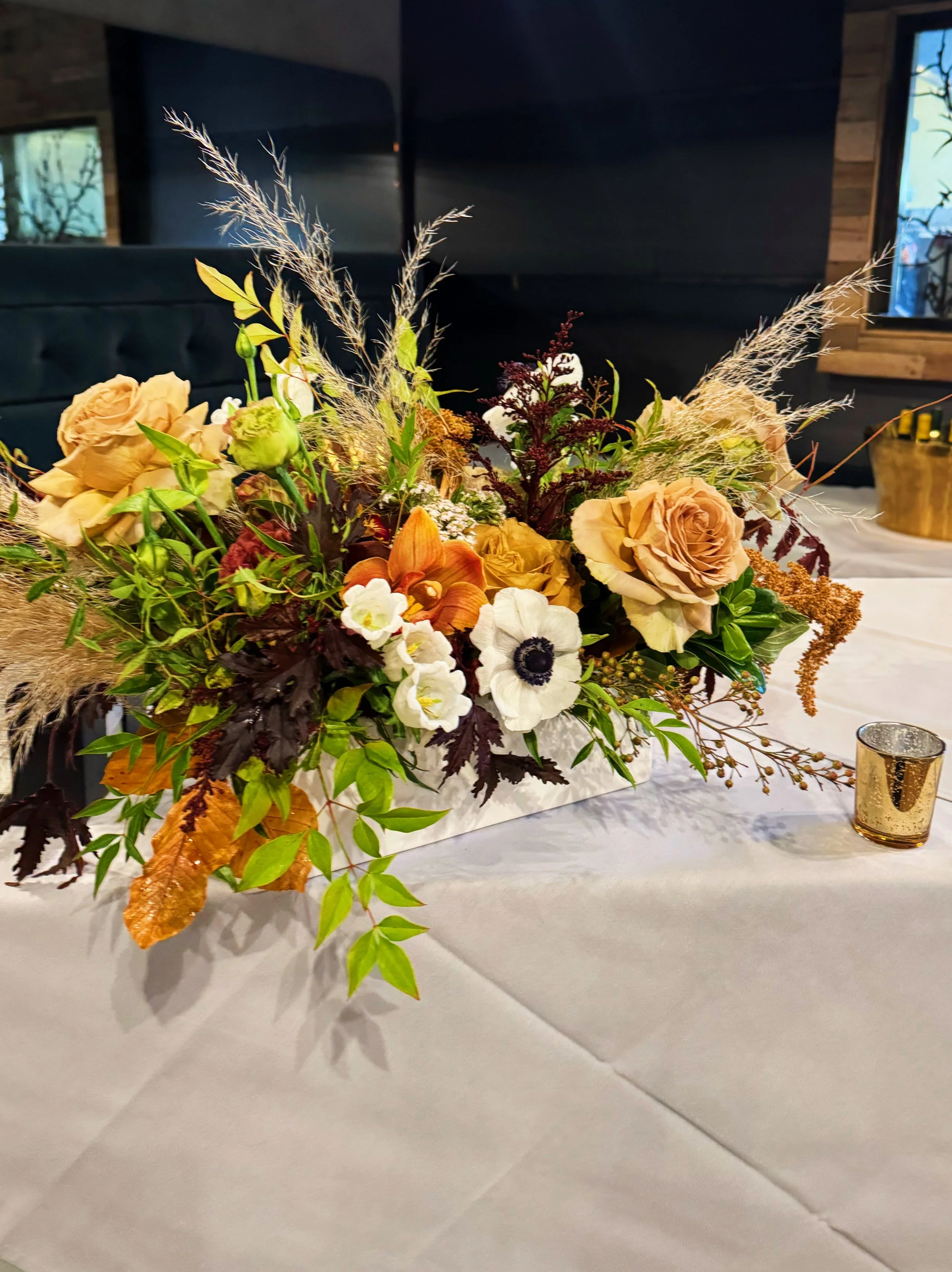 Fall-themed floral centerpiece with roses, anemones, and greenery on a white tablecloth, with a small gold candle holder nearby.