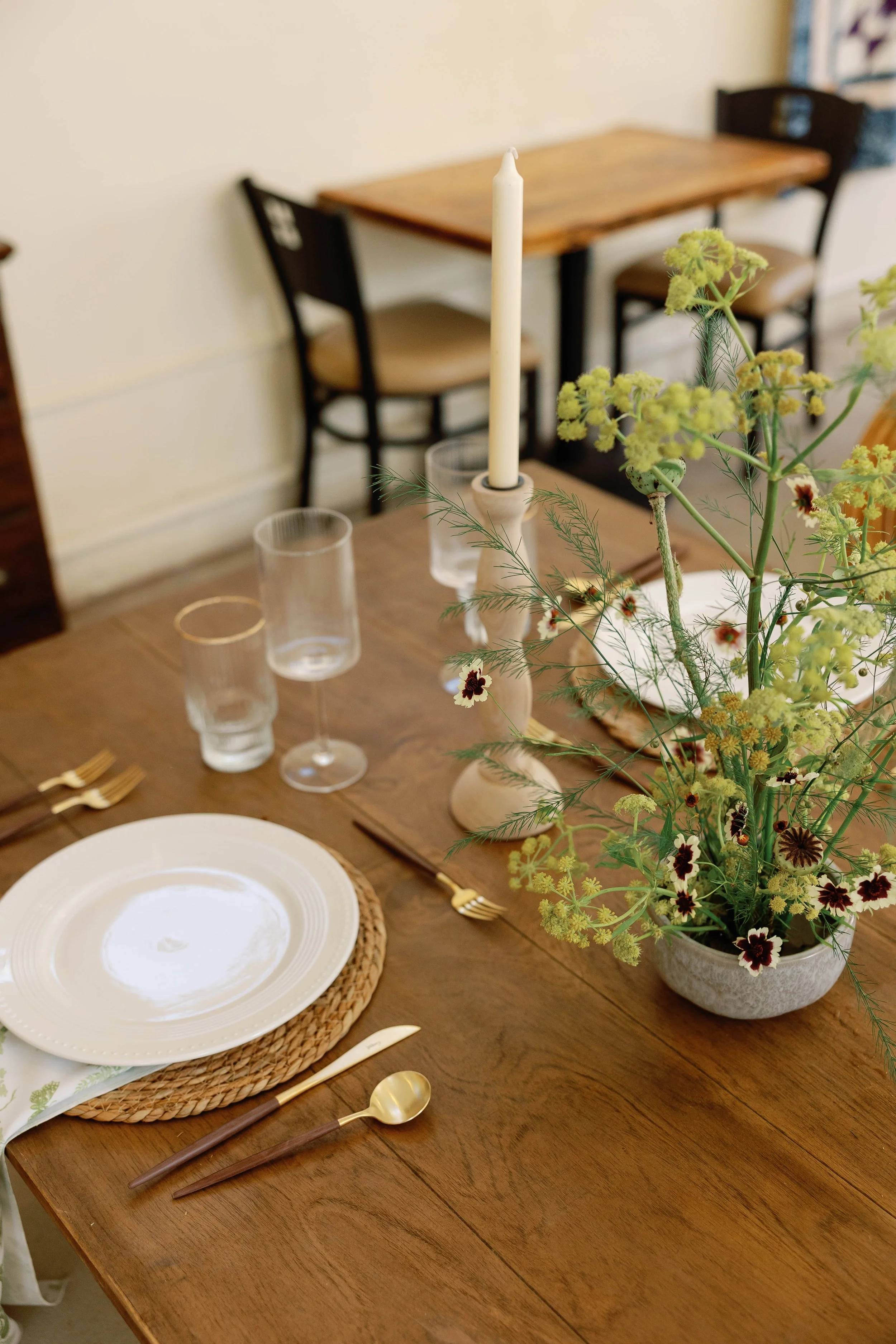 A dining table setup with plates, glasses, gold utensils, a floral centerpiece, and a candle in a holder.