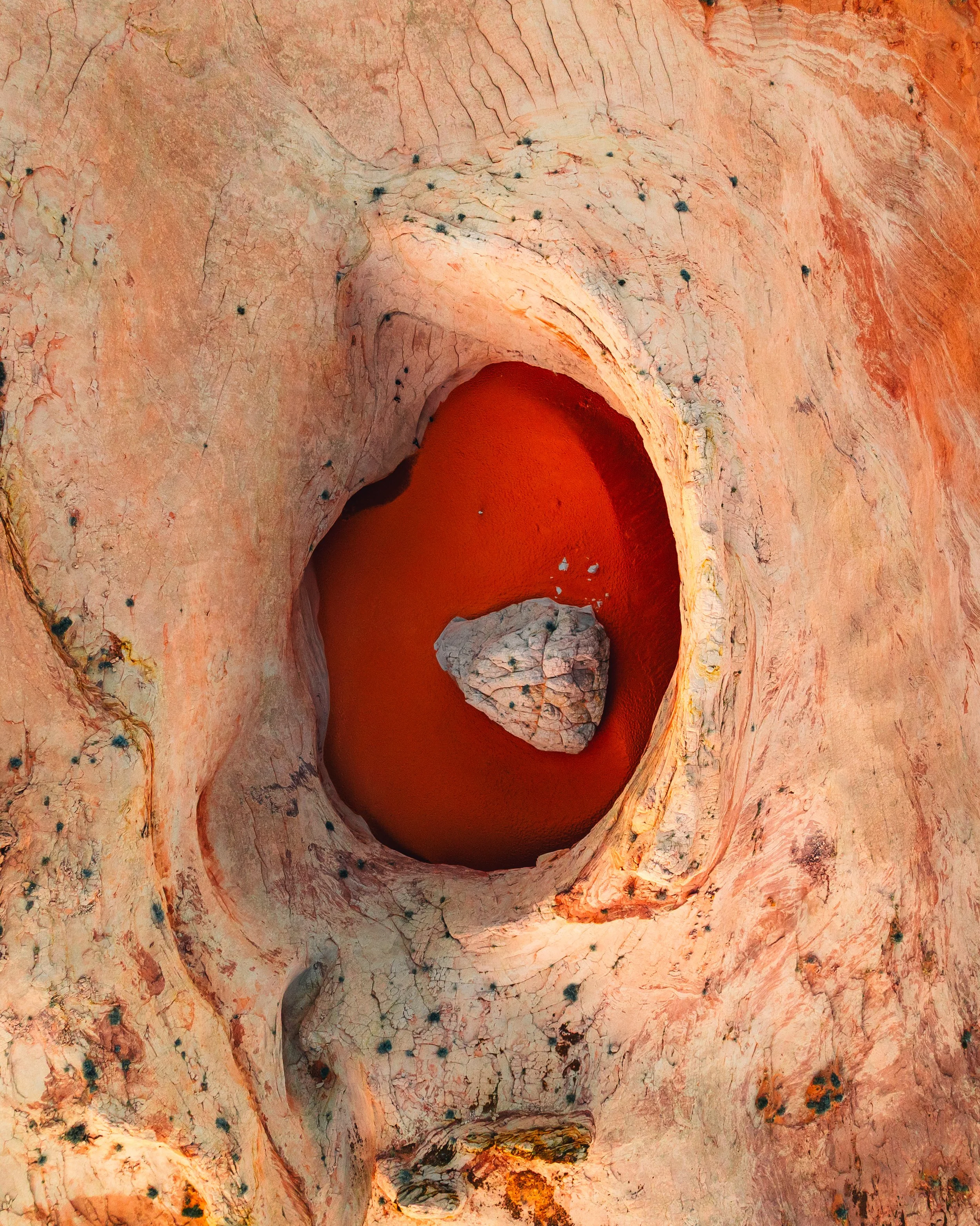 A geology formation showing a deep, oval-shaped hole in pinkish-red sedimentary rock with a large rock at the bottom. The surrounding rock has cracks and black spots.