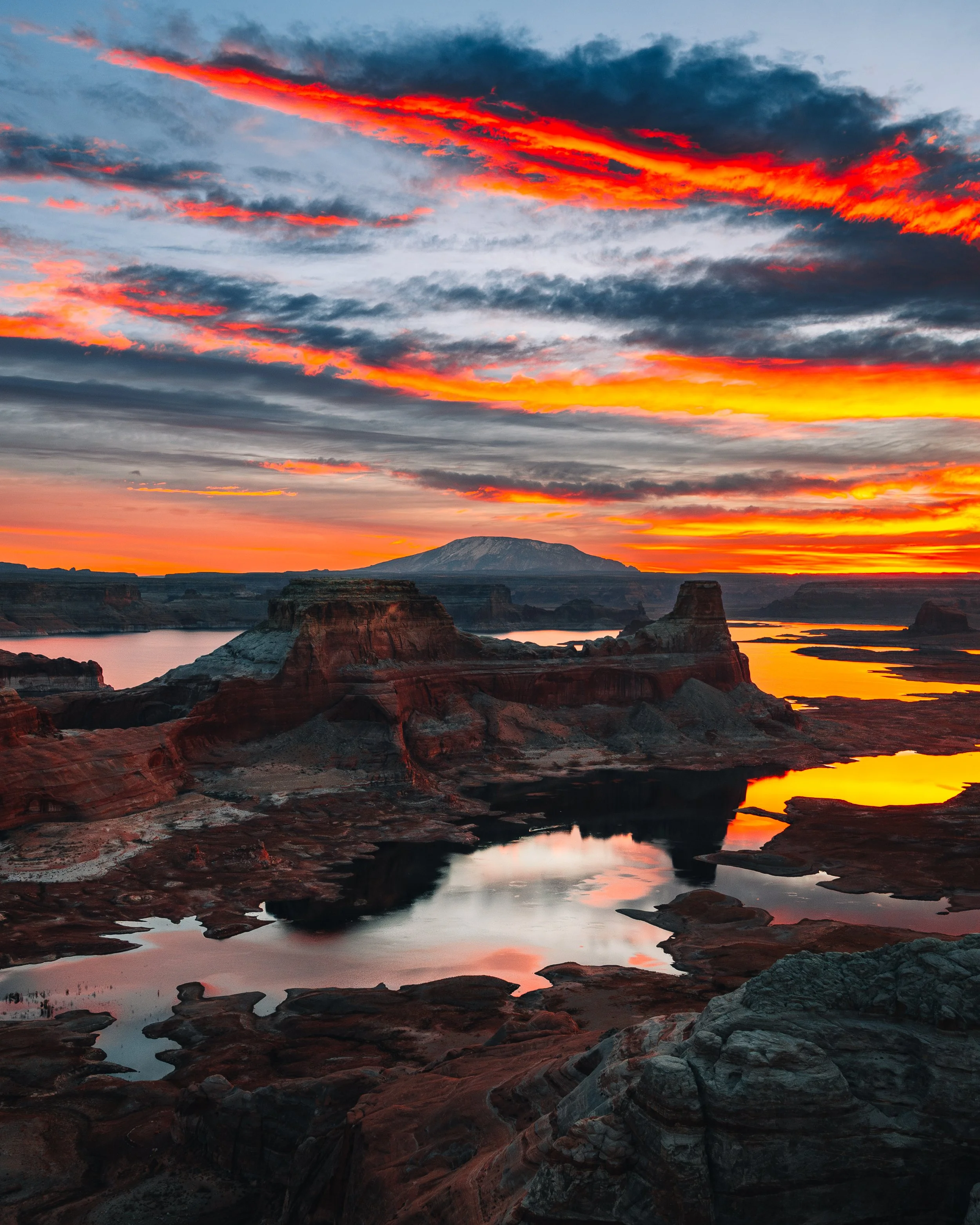 A dramatic sunset over a canyon with layered cliffs, reflecting pools, and a distant mountain, with vibrant orange, red, and blue sky.