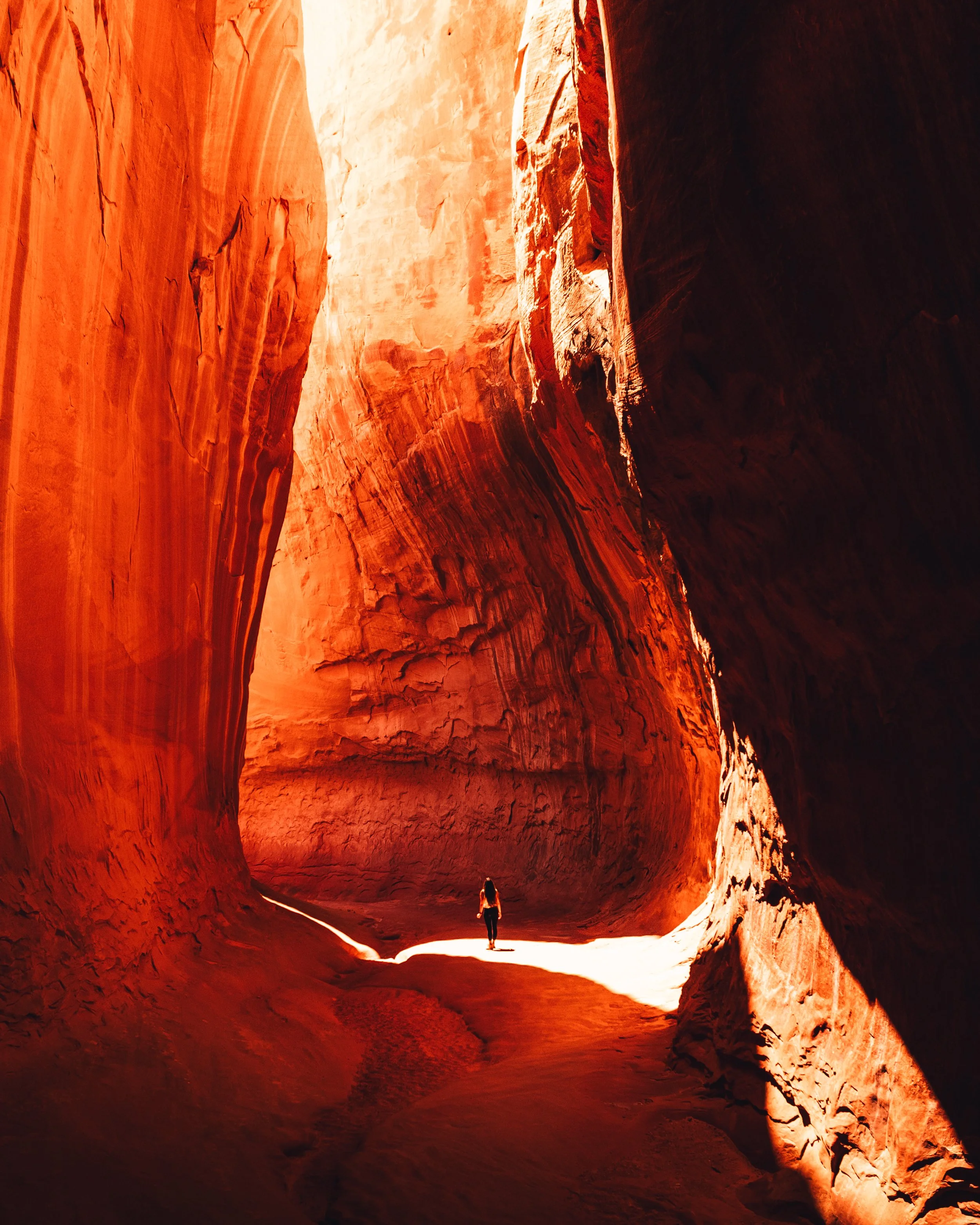 A person walking through a narrow, tall, red and orange carved canyon with sunlight illuminating the walls.