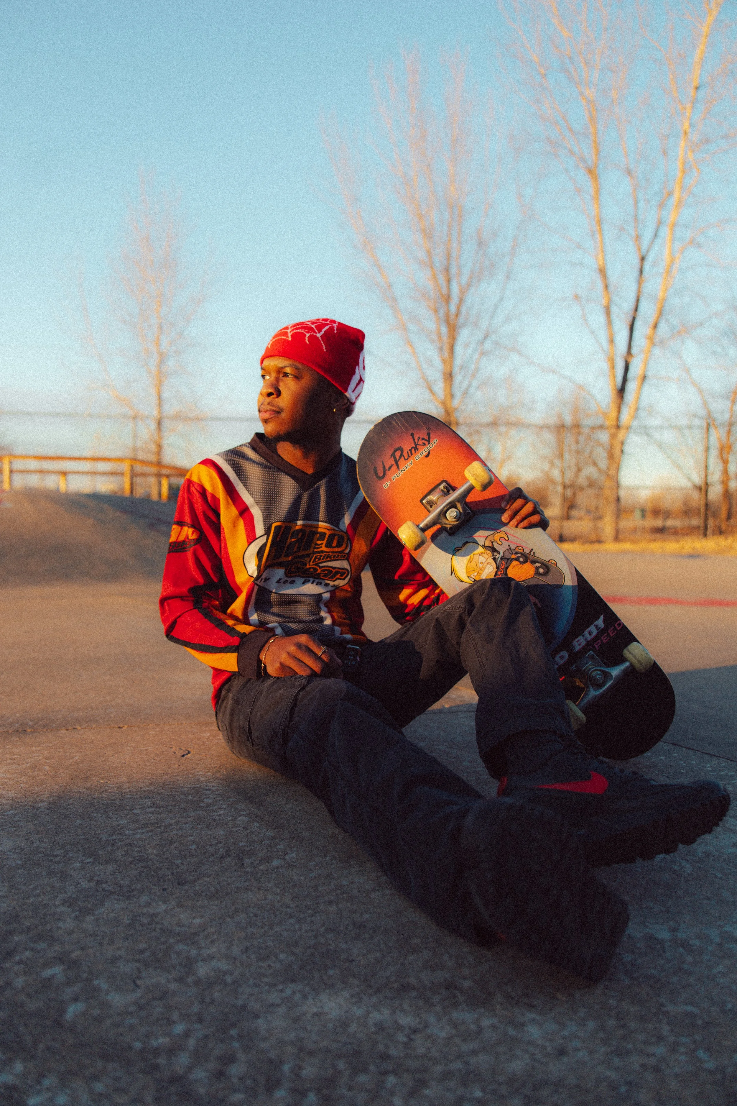 A young man sitting on the ground at a skate park, holding a skateboard, wearing a red beanie and a colorful hockey jersey, with leafless trees in the background during sunset.