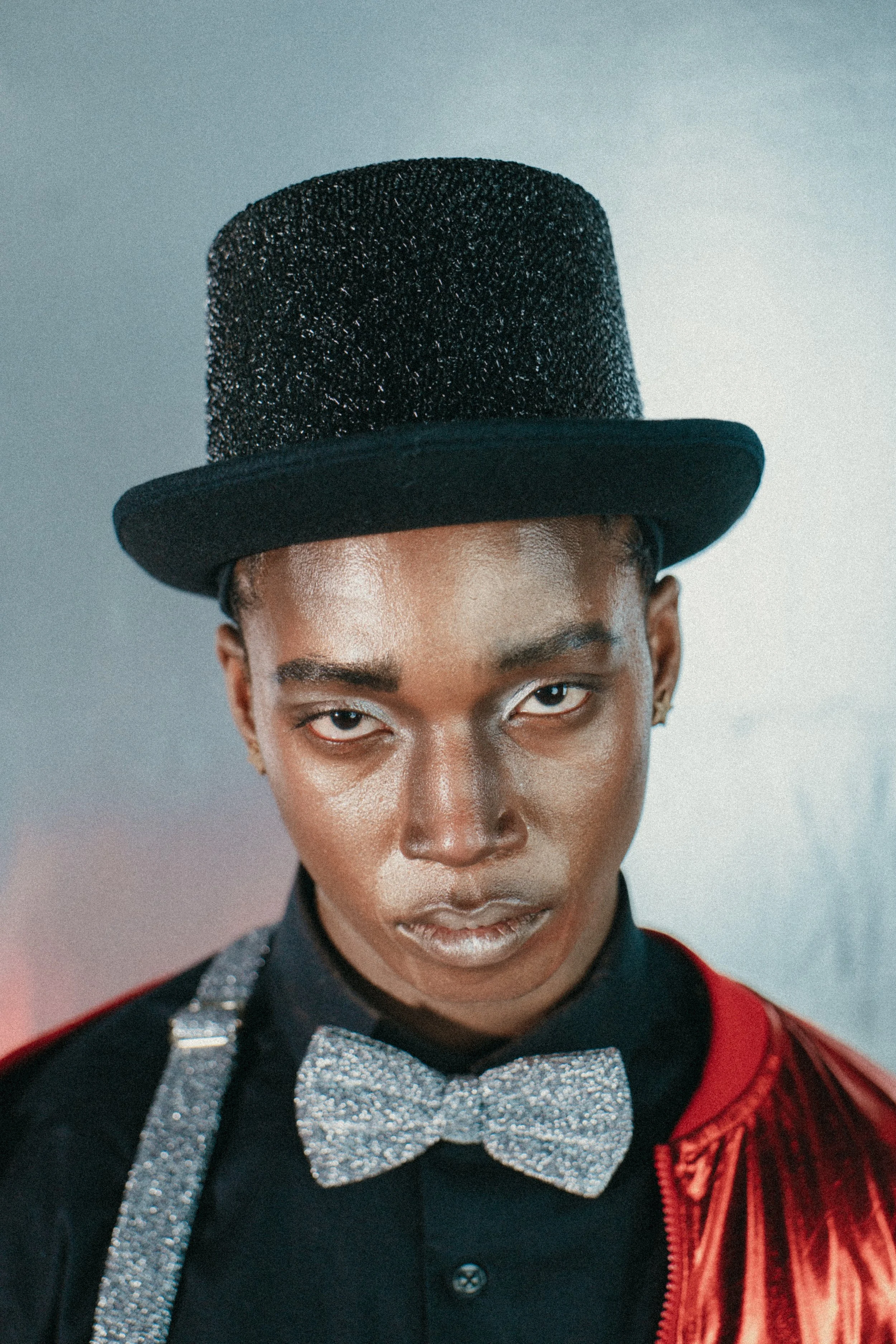 A person wearing a black glittery top hat, a sparkly silver bow tie, and a red jacket with a black shirt, looking directly at the camera with a serious expression.