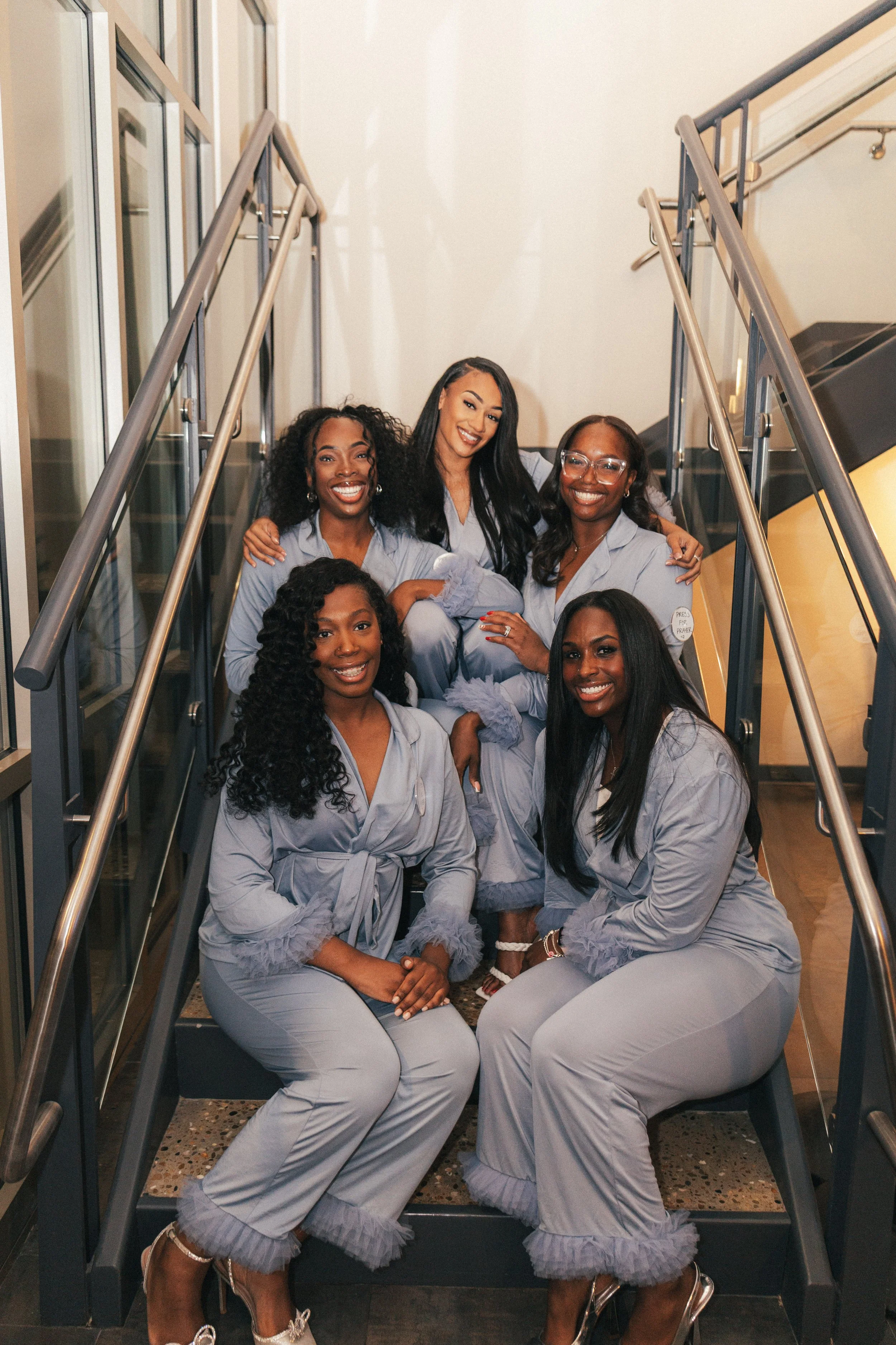 Six women in matching light gray pajamas sitting and standing on a staircase, smiling for a group photo.