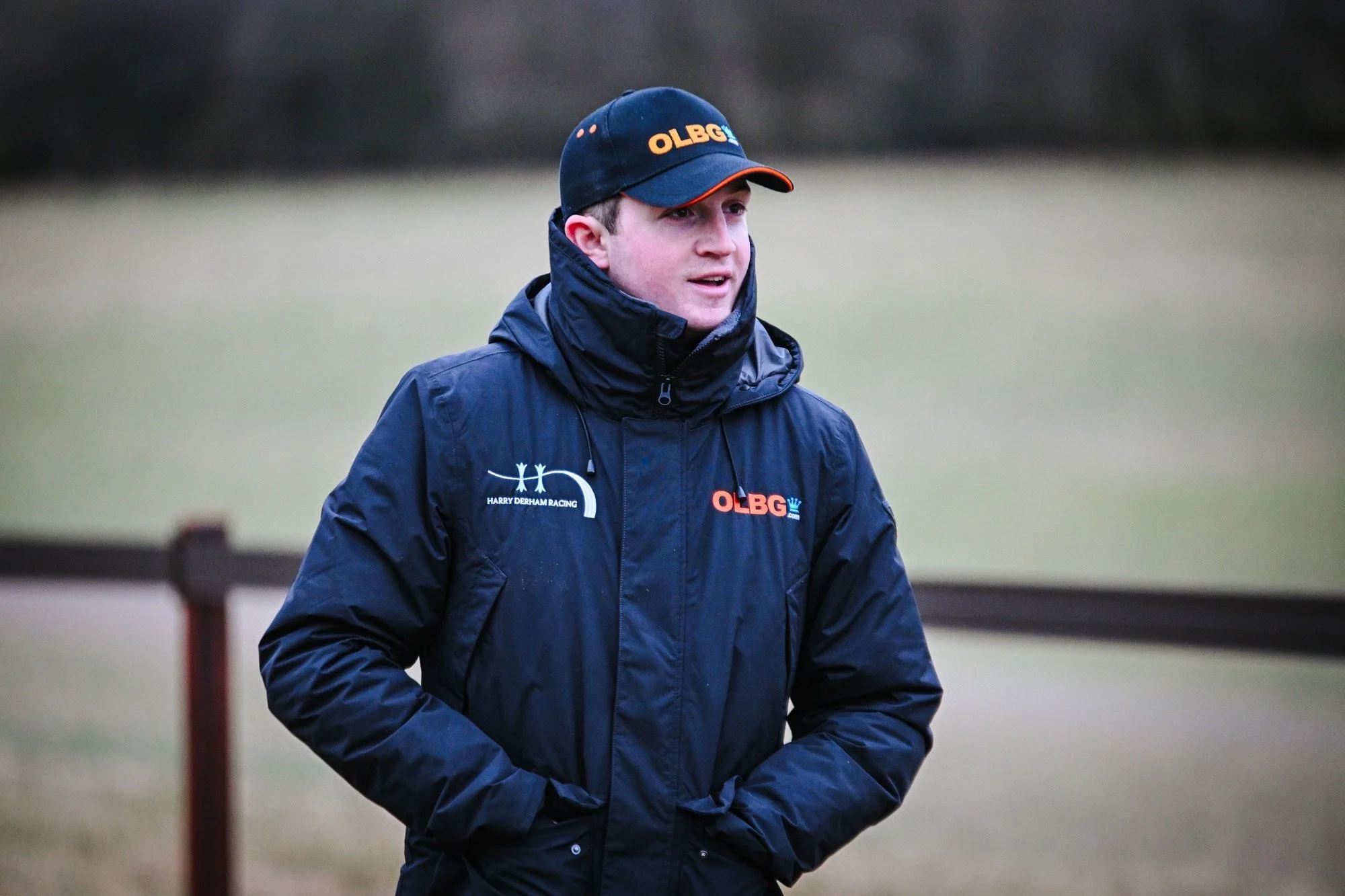 A person standing next to a brown horse wearing a halter, outdoors near a wooden fence. The person is wearing a black jacket and a black cap with logos.