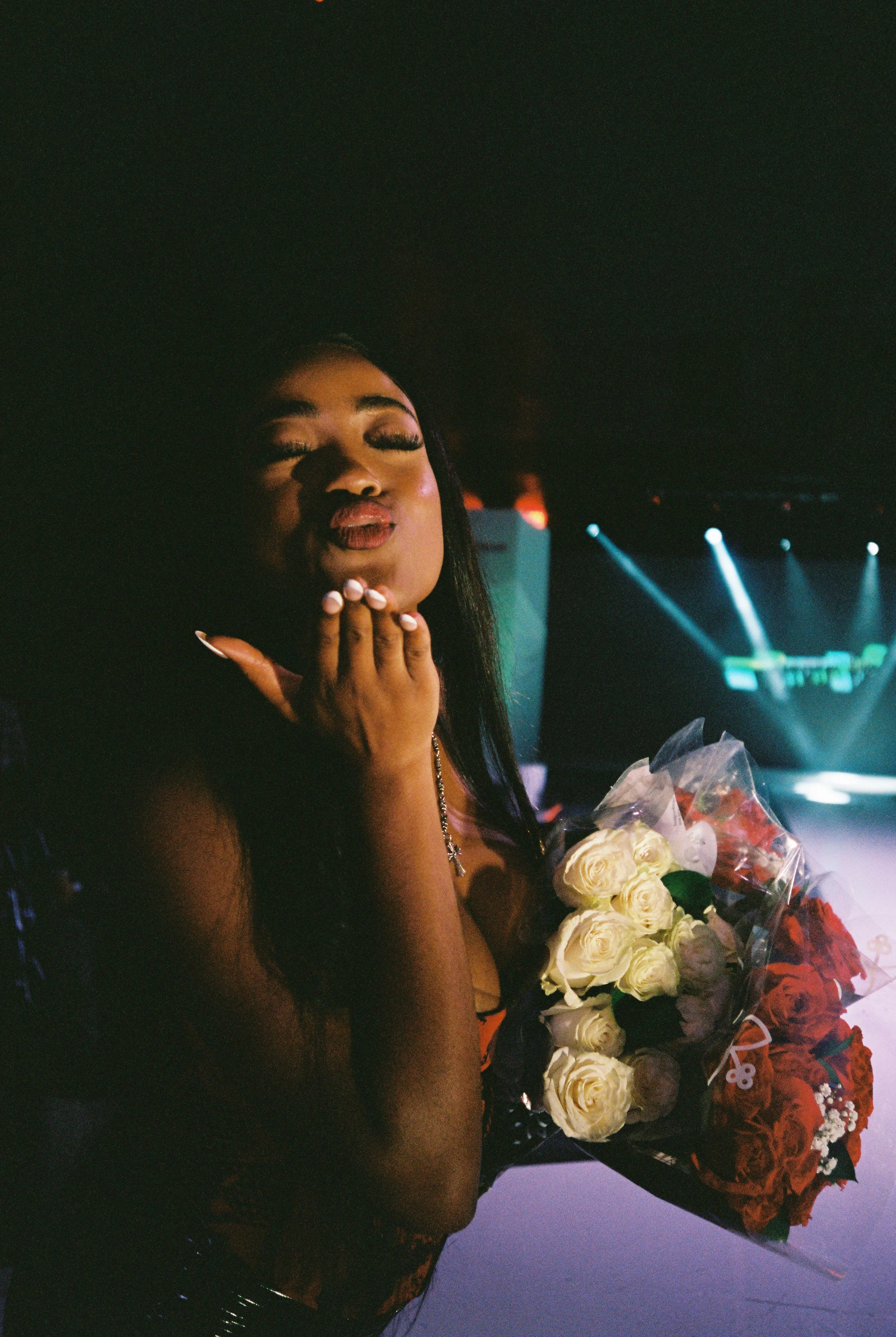 Woman blowing a kiss while holding a bouquet of roses at an event with stage lighting.