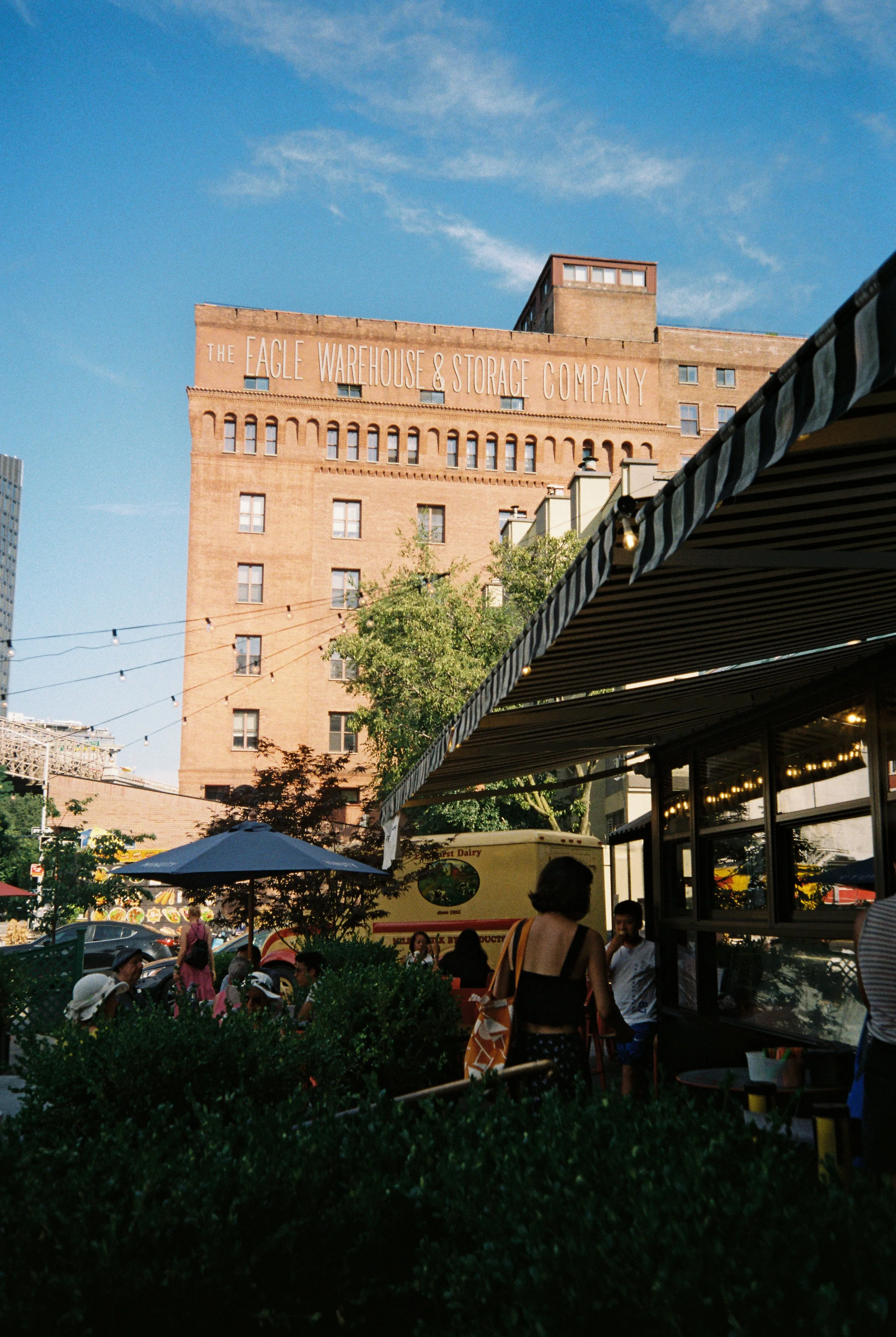 Outdoor cafe scene with people sitting under umbrellas and a view of a historic building labeled 'Eagle Warehouse & Storage Company' in the background, with a clear blue sky.