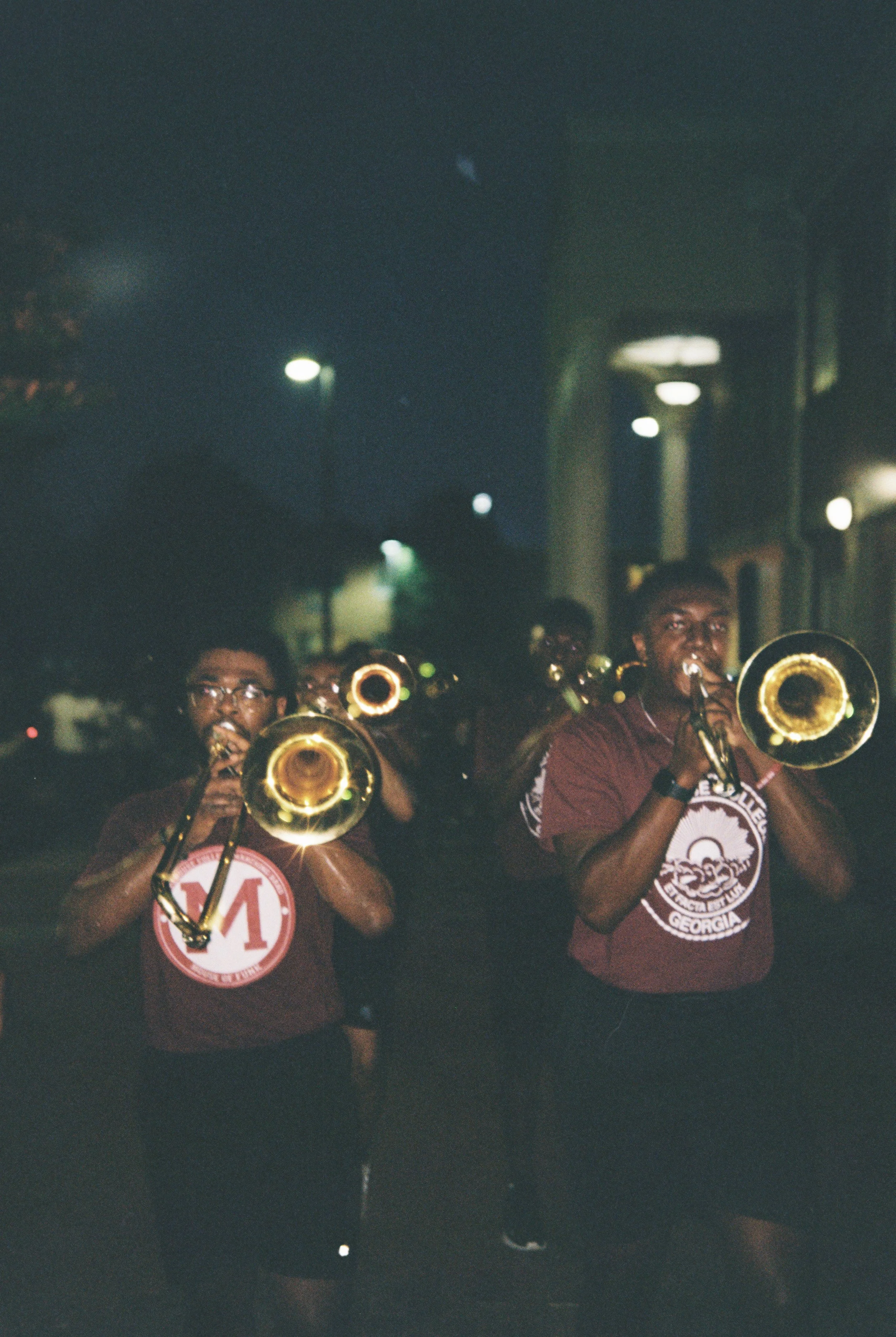 Marching band members playing trombones at night, wearing maroon t-shirts, in a street setting.