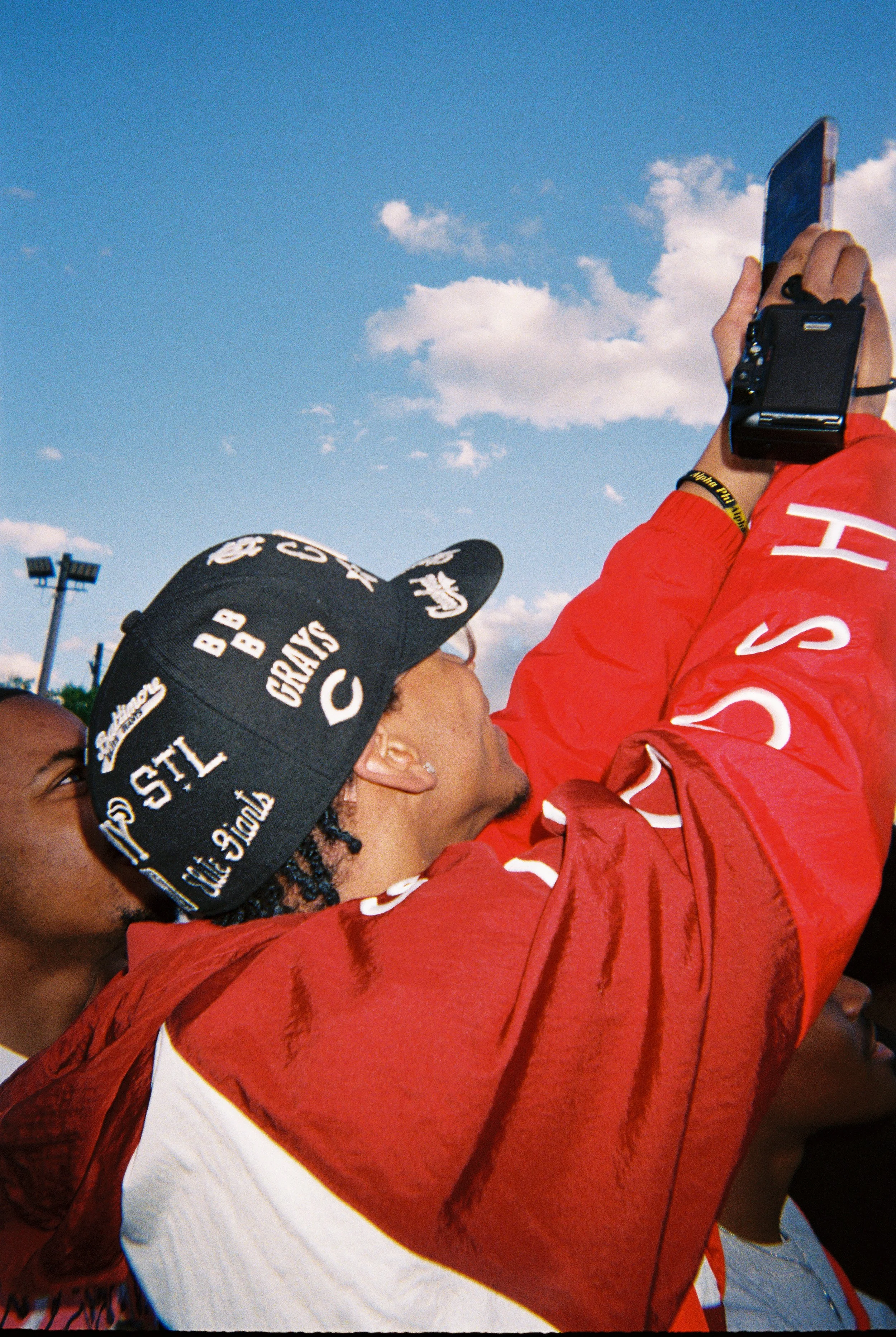 Person wearing a black cap with embroidered logos taking a photo with a camera, wearing a red jacket, under a cloudy blue sky.