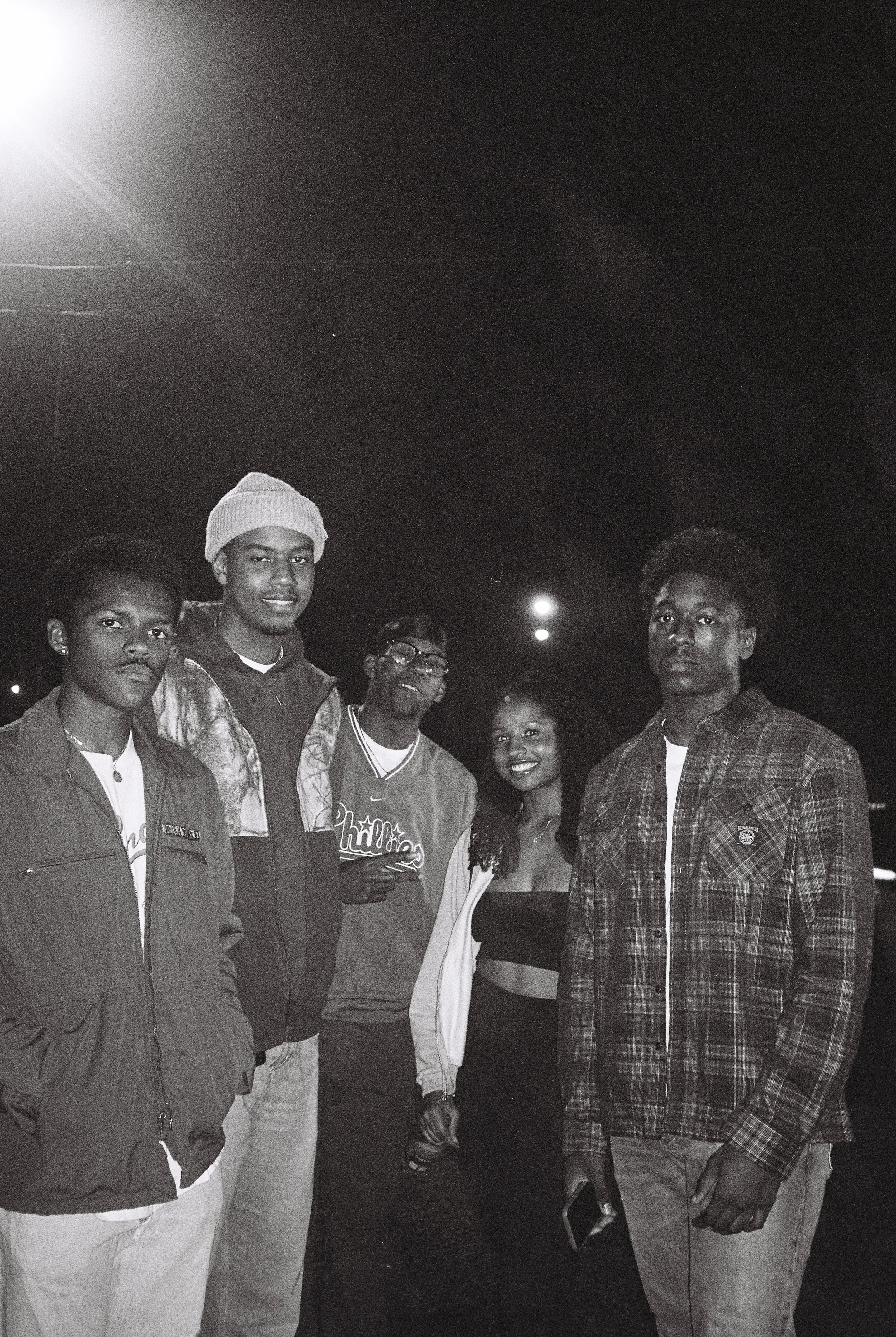 Group of five people posing outdoors at night, three men wearing jackets and one wearing a beanie, one woman smiling, another man in a plaid shirt holding a phone.