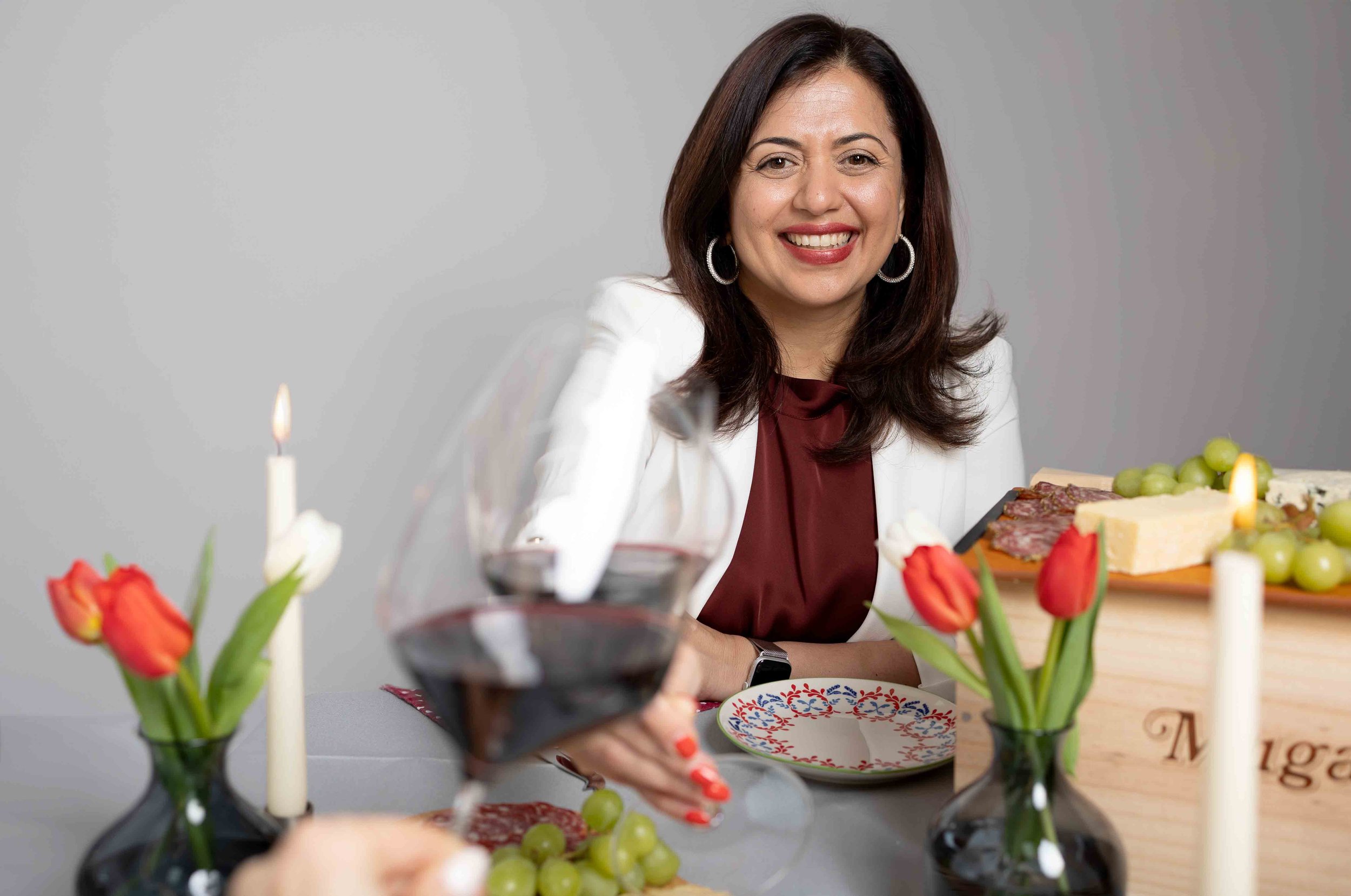 A smiling woman with dark brown hair wearing a white blazer and red blouse, sitting at a table decorated with vases of red tulips, candles, and a glass of red wine, with a cheese and meat platter, grapes, and a wooden box of wine in front of her.