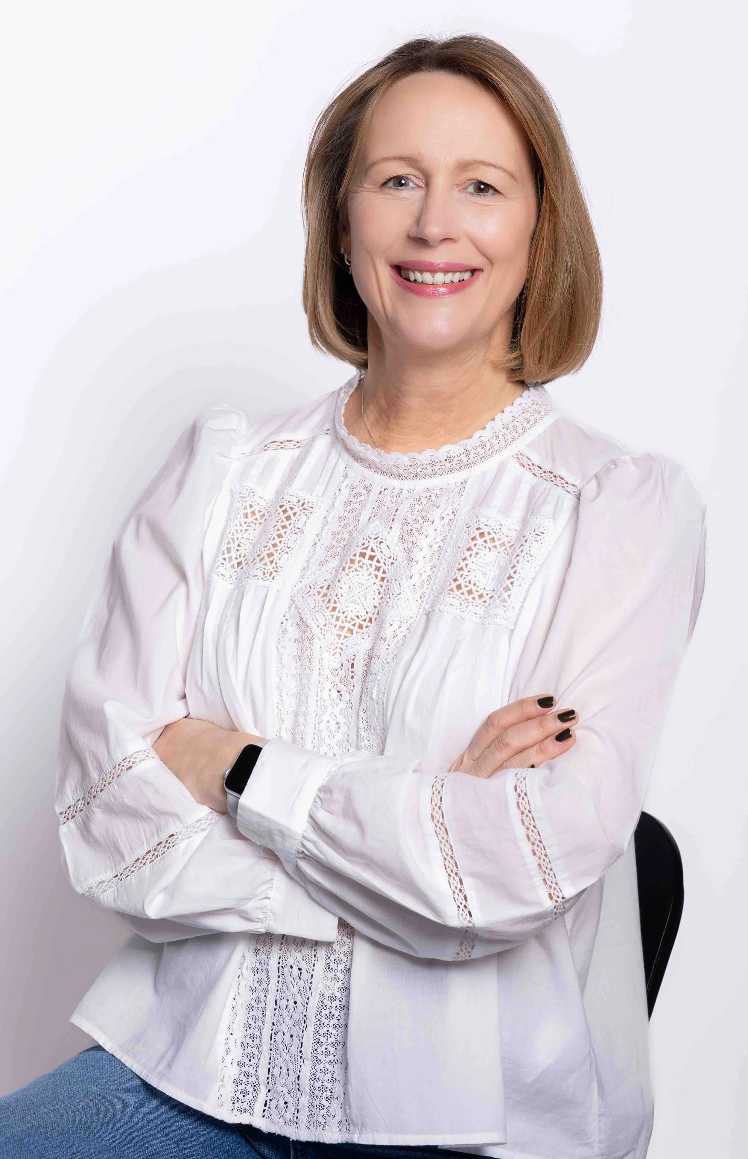 A woman with red hair smiling, dressed in a white embroidered blouse, sitting with crossed arms against an off-white background.