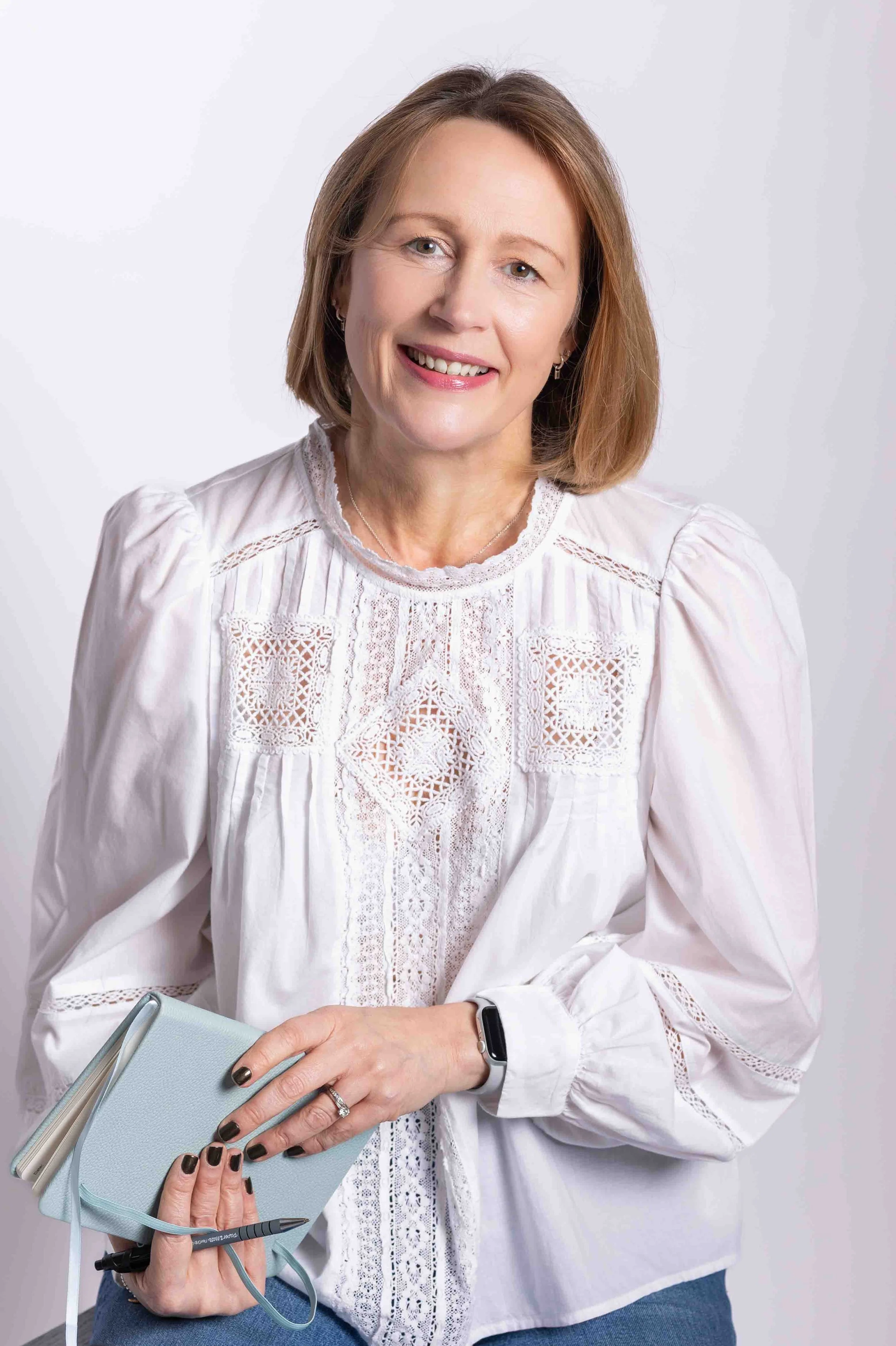 A woman with light brown hair smiling, wearing a white embroidered blouse, holding a light blue notebook and a black pen, against a plain light background.