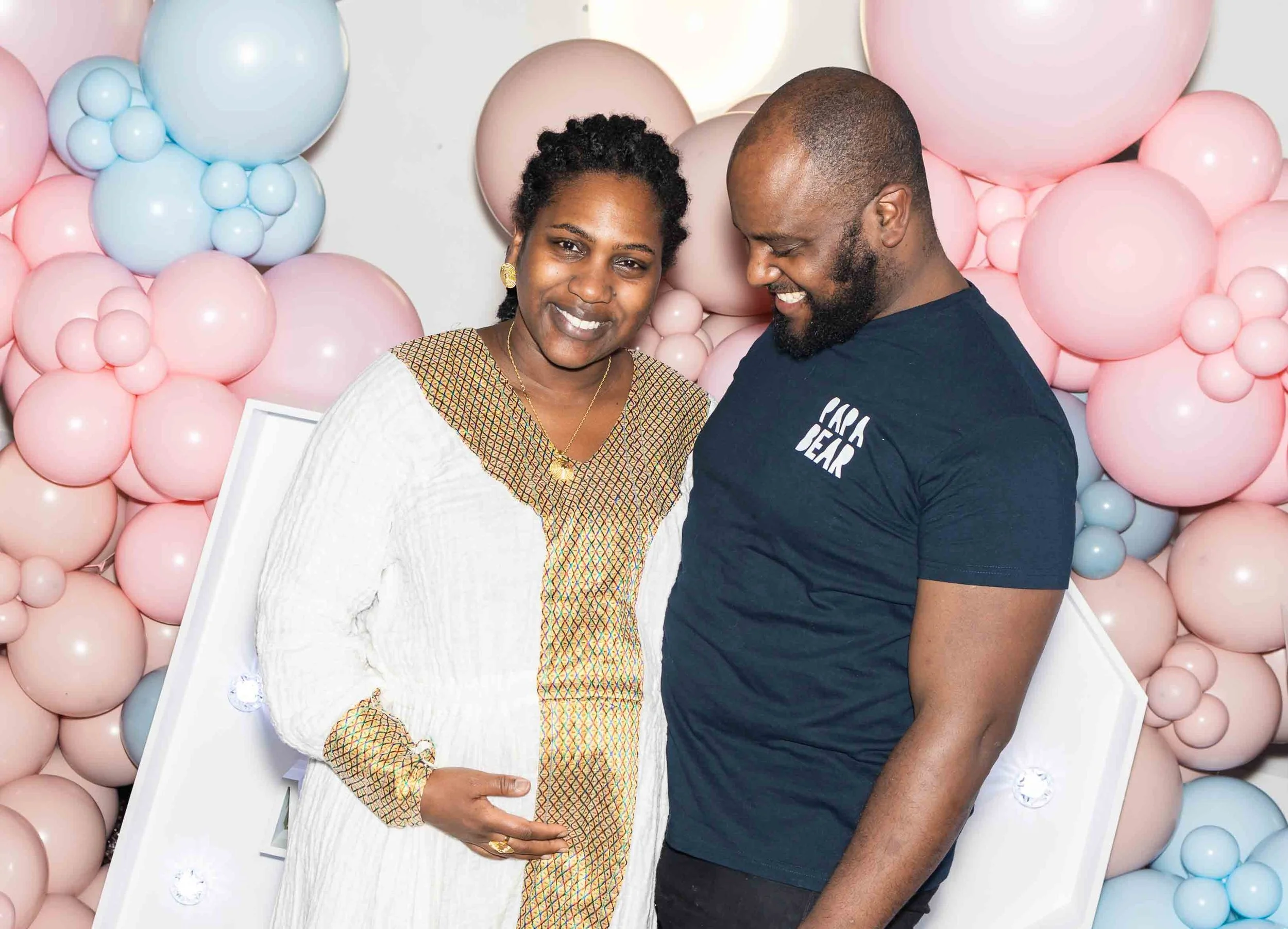 A pregnant woman and a man smiling together in front of pink and blue balloon decorations, with the woman gently resting her hand on her belly.