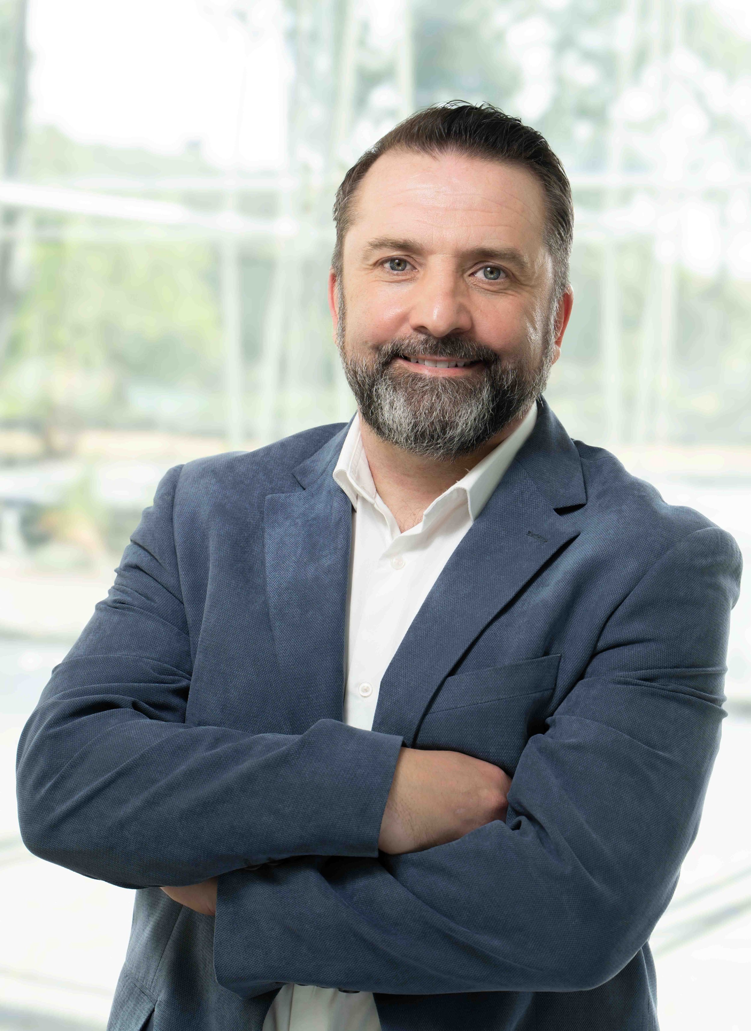 A man with dark hair and a beard, smiling, wearing a white shirt and a blue blazer, standing with arms crossed in front of a bright window with a blurry outdoor background.