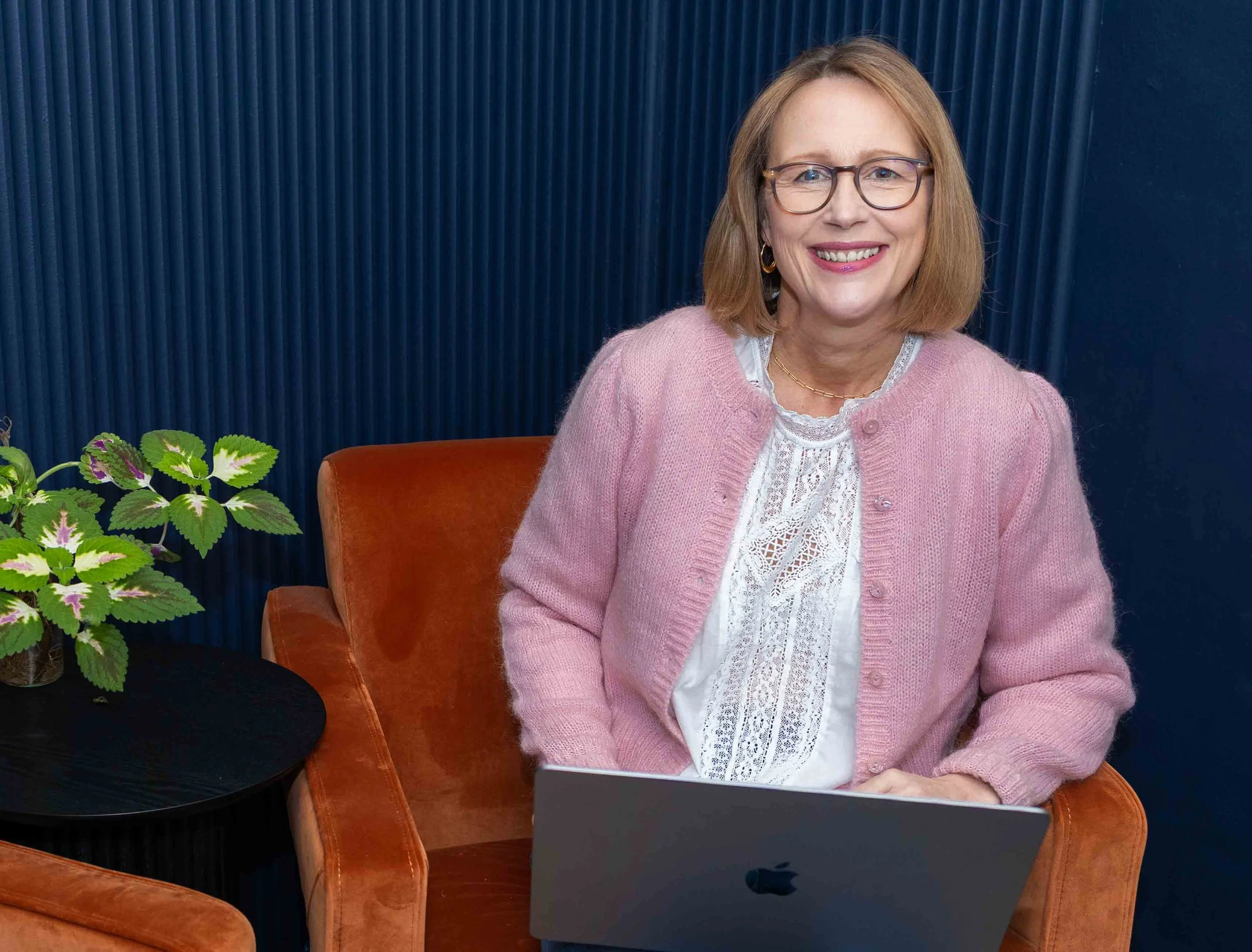 A woman with glasses, blonde hair, and a pink cardigan sitting on an orange velvet armchair, smiling at the camera with a laptop on her lap. There is a green plant with variegated leaves on a black side table next to her, and a dark blue textured wal