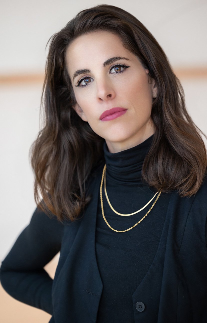 A woman with shoulder-length brown hair, wearing a black turtleneck and gold layered necklaces, looking directly at the camera with a neutral expression.