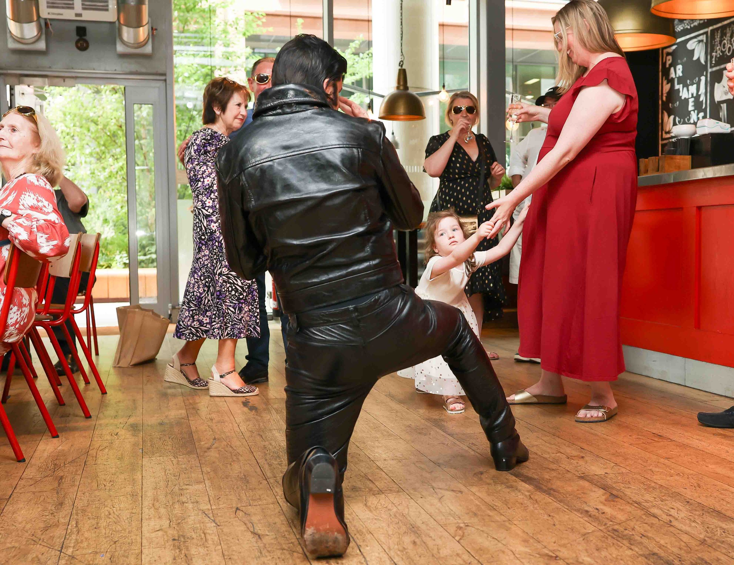 A person dressed as Elvis Presley kneeling and singing to a young girl dressed in white at a social gathering in a restaurant. Other people are standing and sitting around, some with drinks, with large windows and wooden flooring in the background.