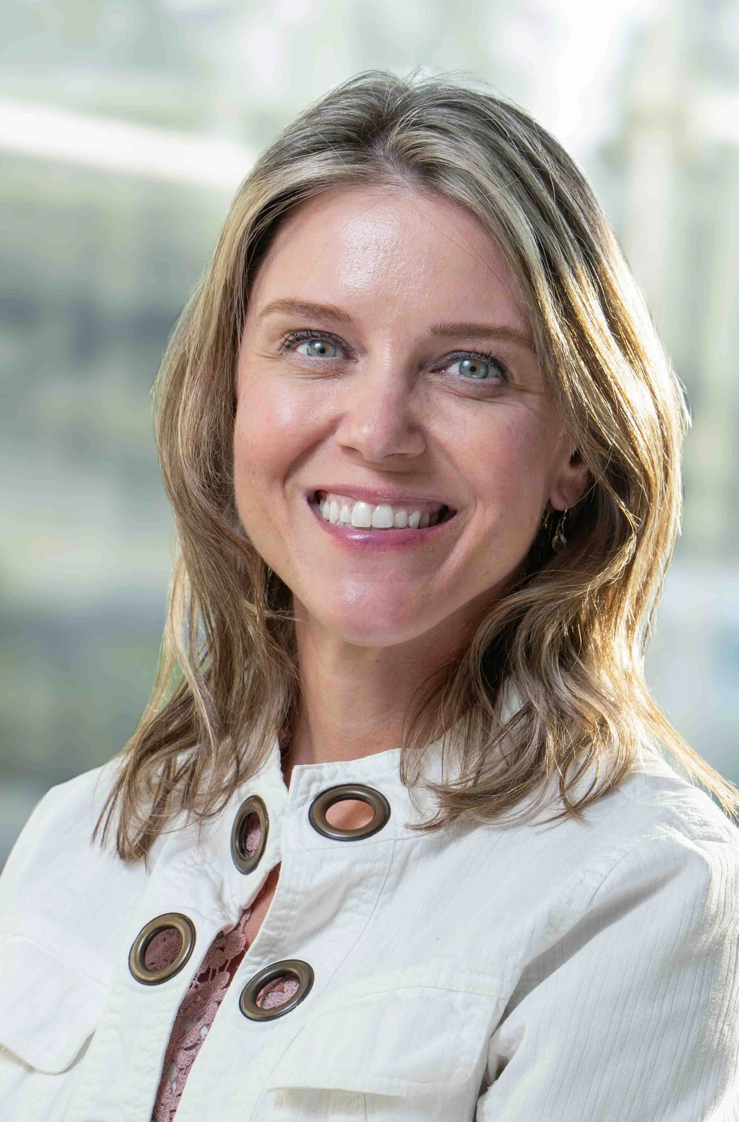 A smiling woman with shoulder-length light brown hair, wearing a white jacket with large buttons, standing indoors with a blurred background.