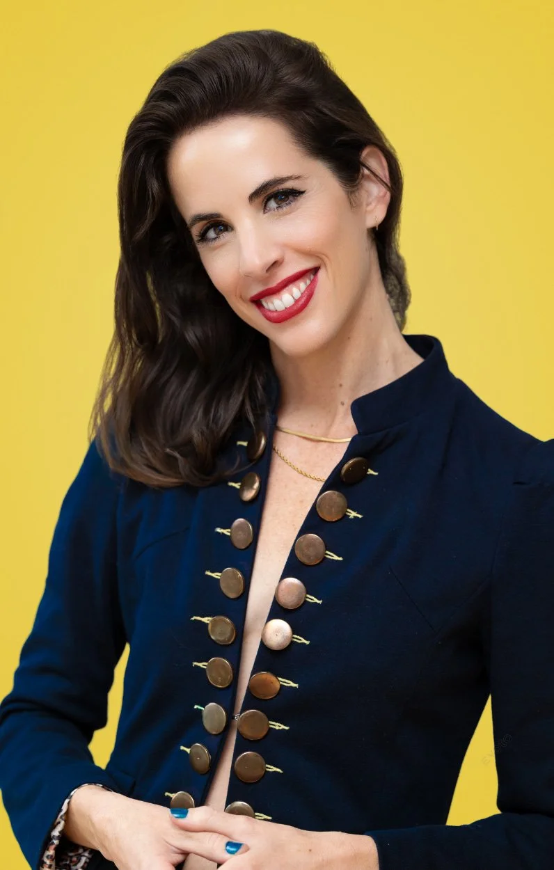 A woman with dark brown hair, wearing red lipstick, a navy blue top with large buttons, and a gold necklace, smiling against a yellow background.