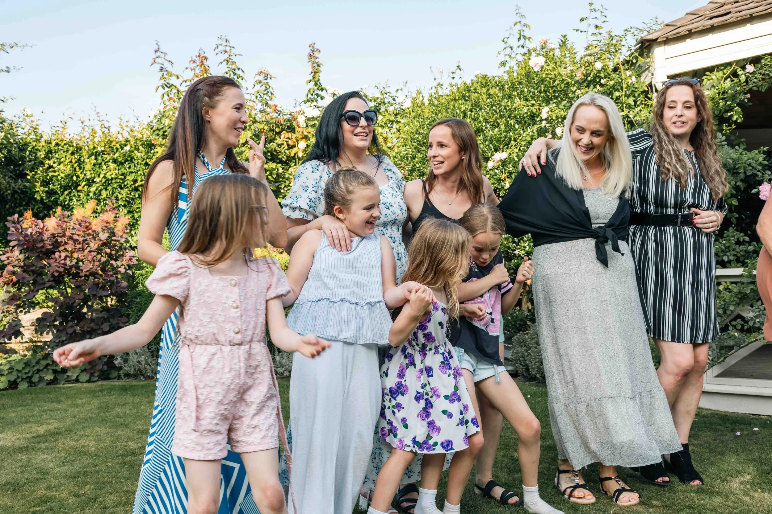 Group of women and children enjoying an outdoor gathering in a garden, smiling and interacting with each other.