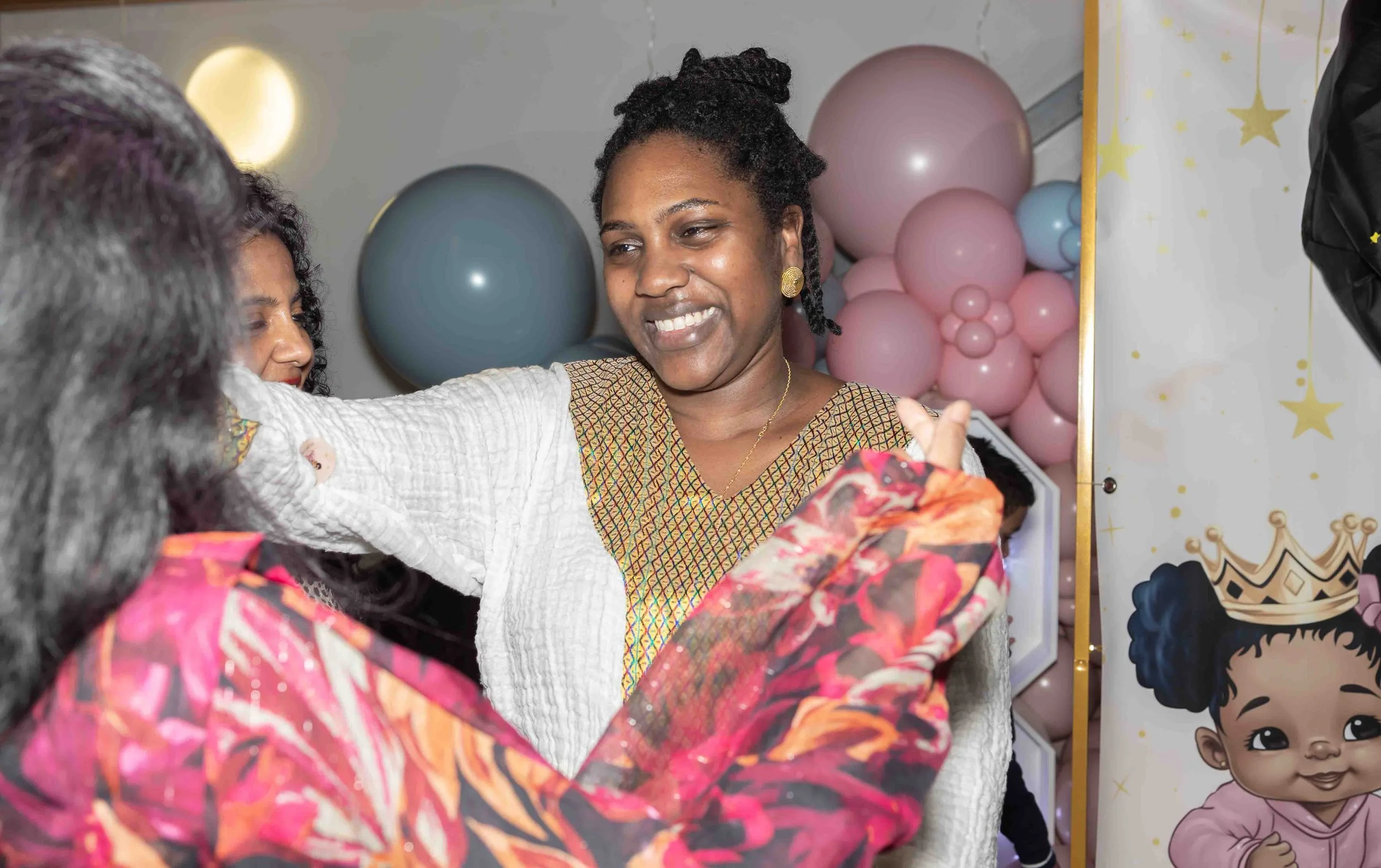 A woman with dark hair styled in braids wears a colorful dress with a white jacket, smiling as she looks at someone taking a photo. Pink and blue balloons and a decorative banner with an illustration of a girl with curly hair and crown are in the bac