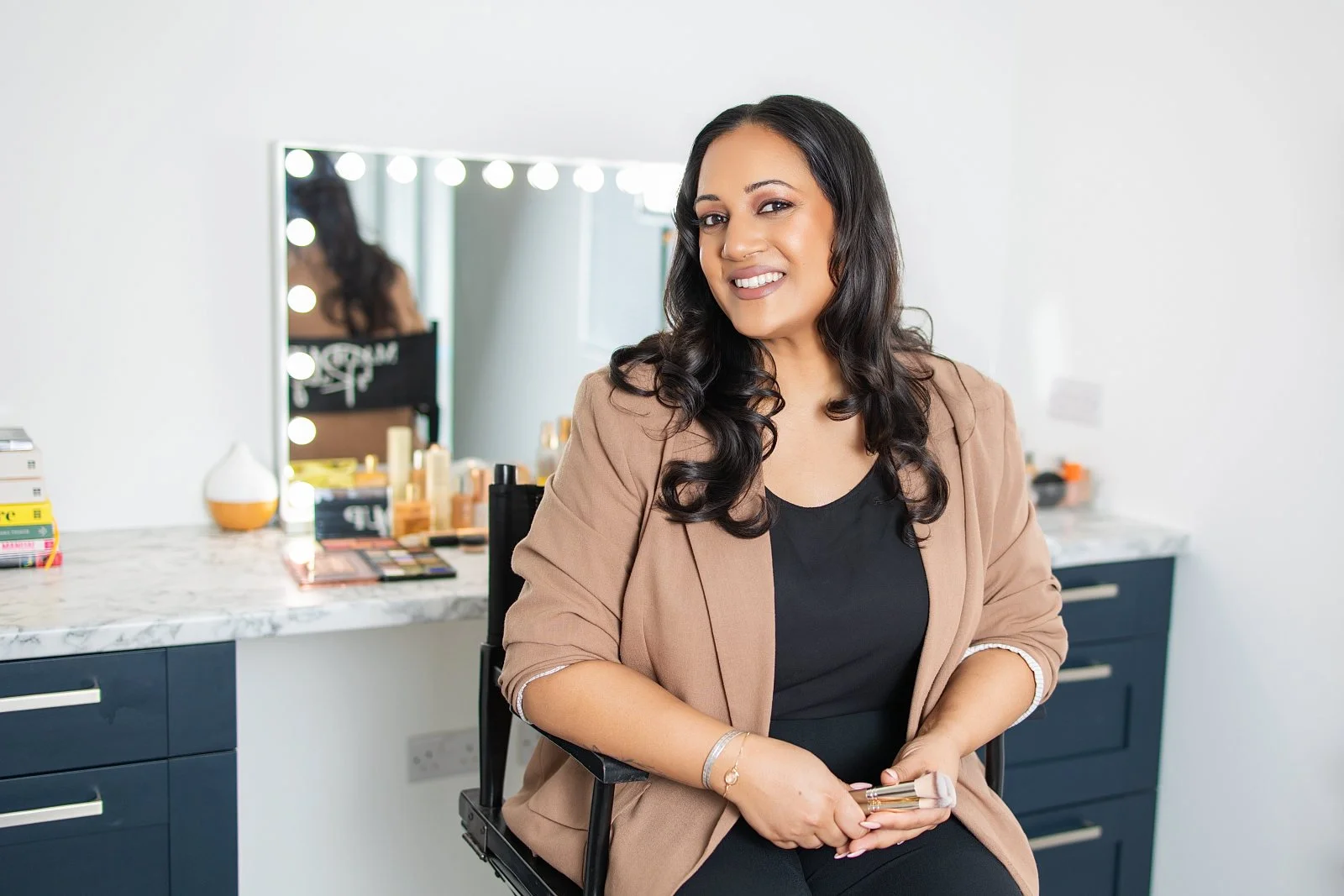 A woman with dark, wavy hair, wearing a black top and a beige blazer, sitting in front of a mirror with round lights, smiling and holding a makeup brush. The background features a makeup station with various beauty products.