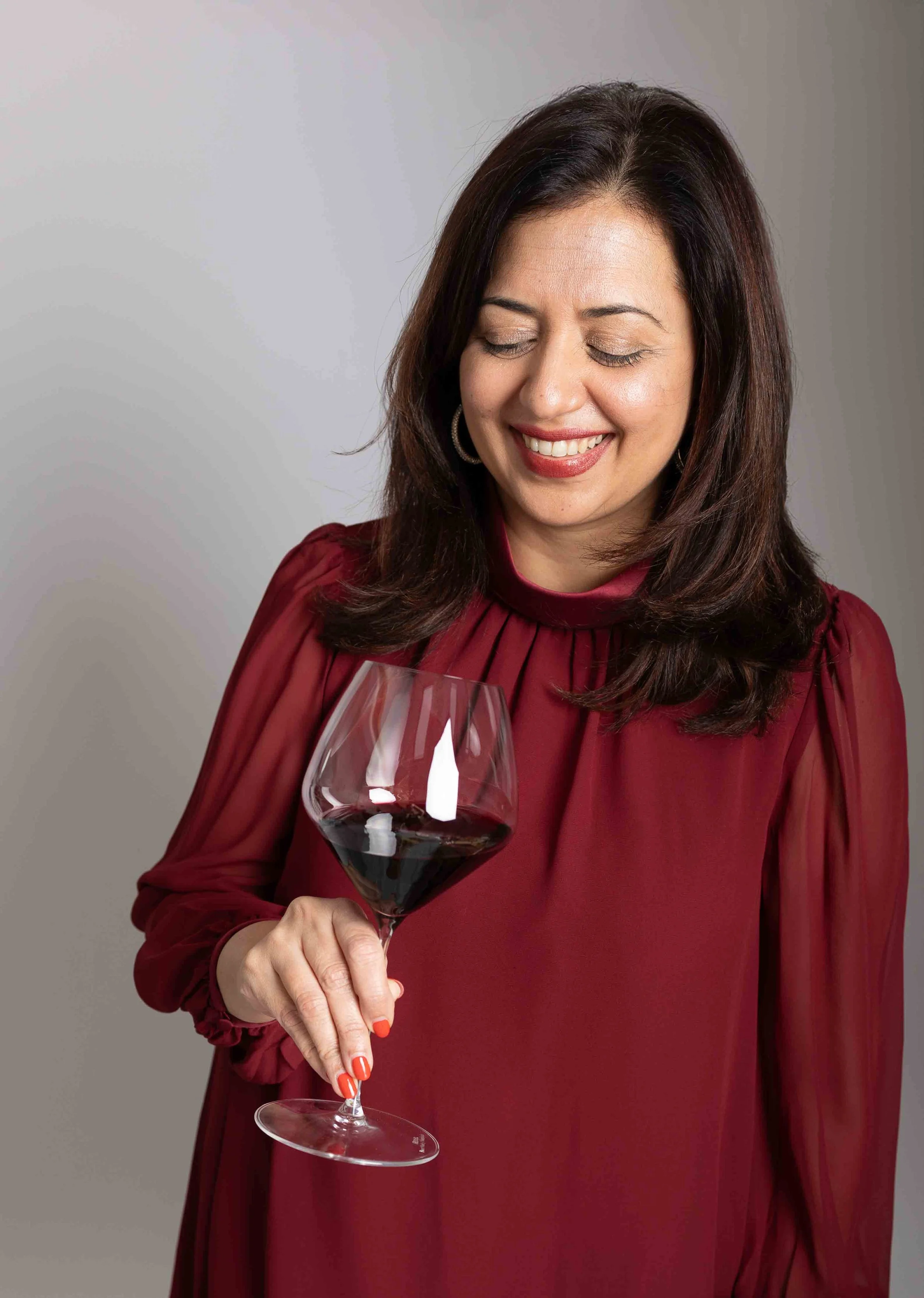 A woman with dark hair, wearing a red dress, holding a glass of red wine and smiling with her eyes closed.