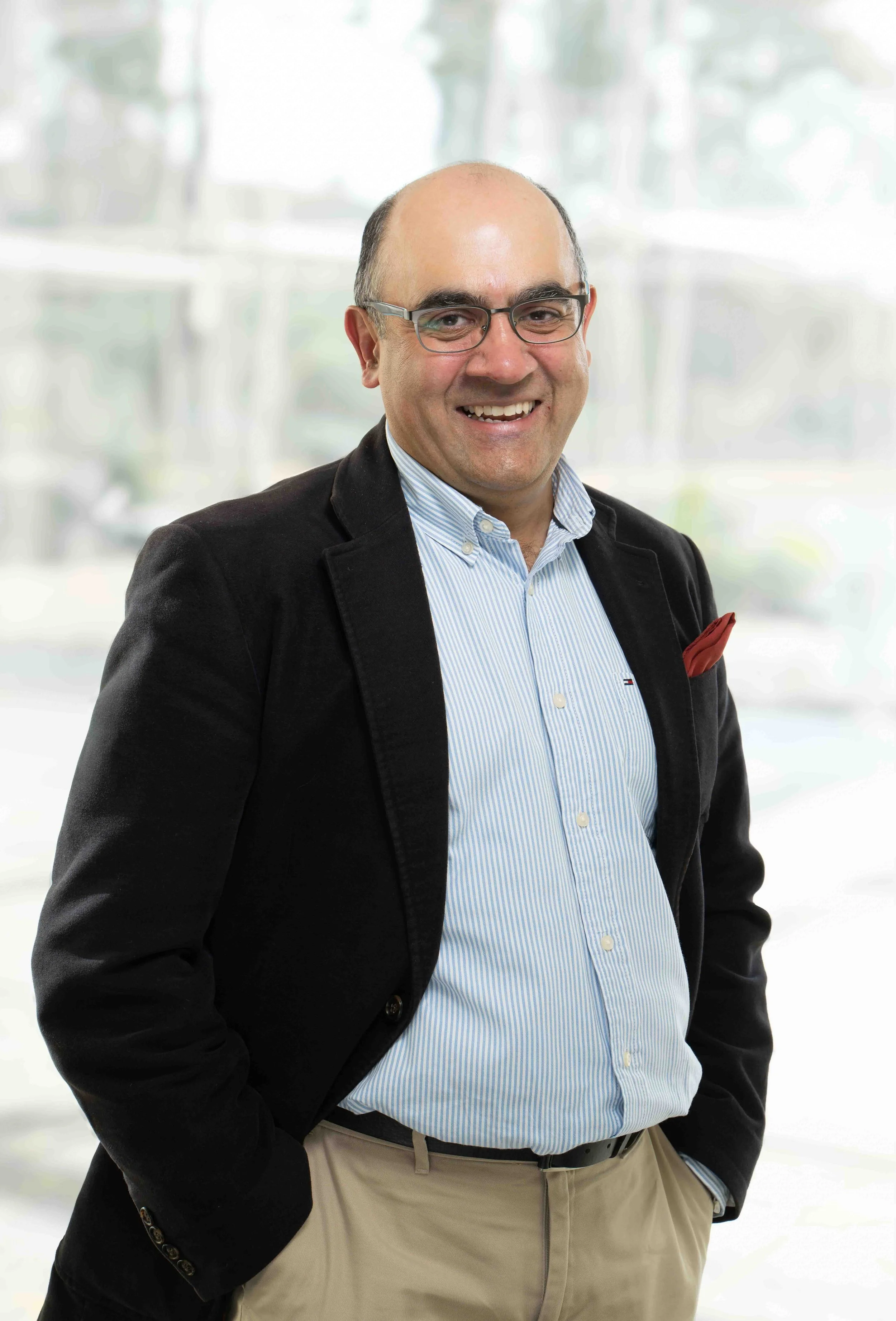 A man with glasses, wearing a dark blazer, light blue striped shirt, and beige pants, standing with his hands in his pockets in front of a bright, blurred background.