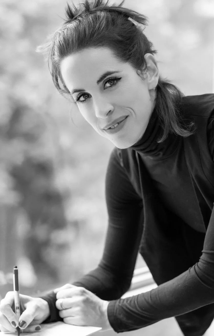Black and white photo of a woman with dark hair and light eyes, wearing a turtleneck and a jacket, smiling and holding a pen while writing on paper outdoors.