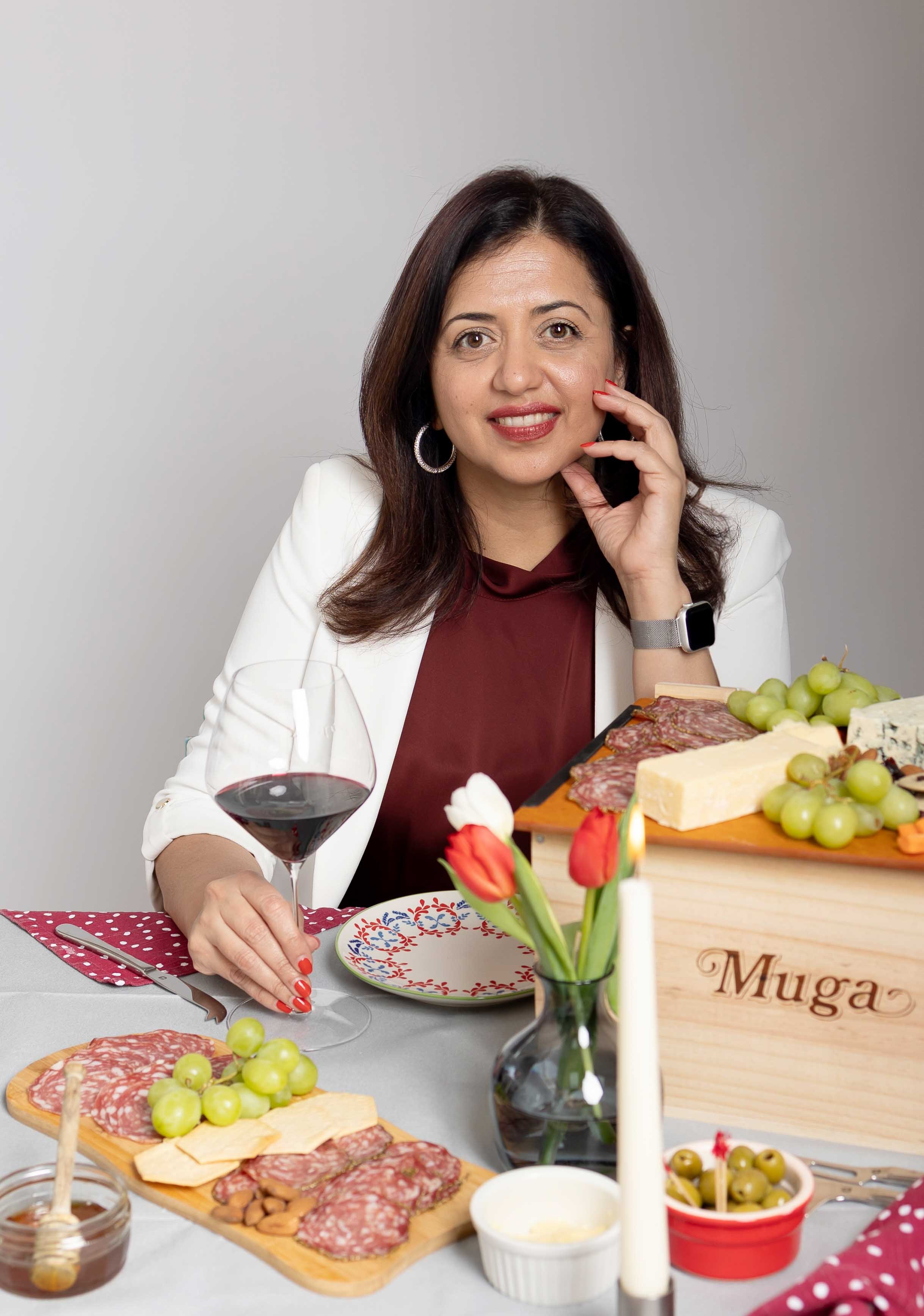 Woman with dark brown hair and hoop earrings sitting at a table with a glass of red wine, cheese, grapes, cold cuts, and a cheese box labeled 'Muga'