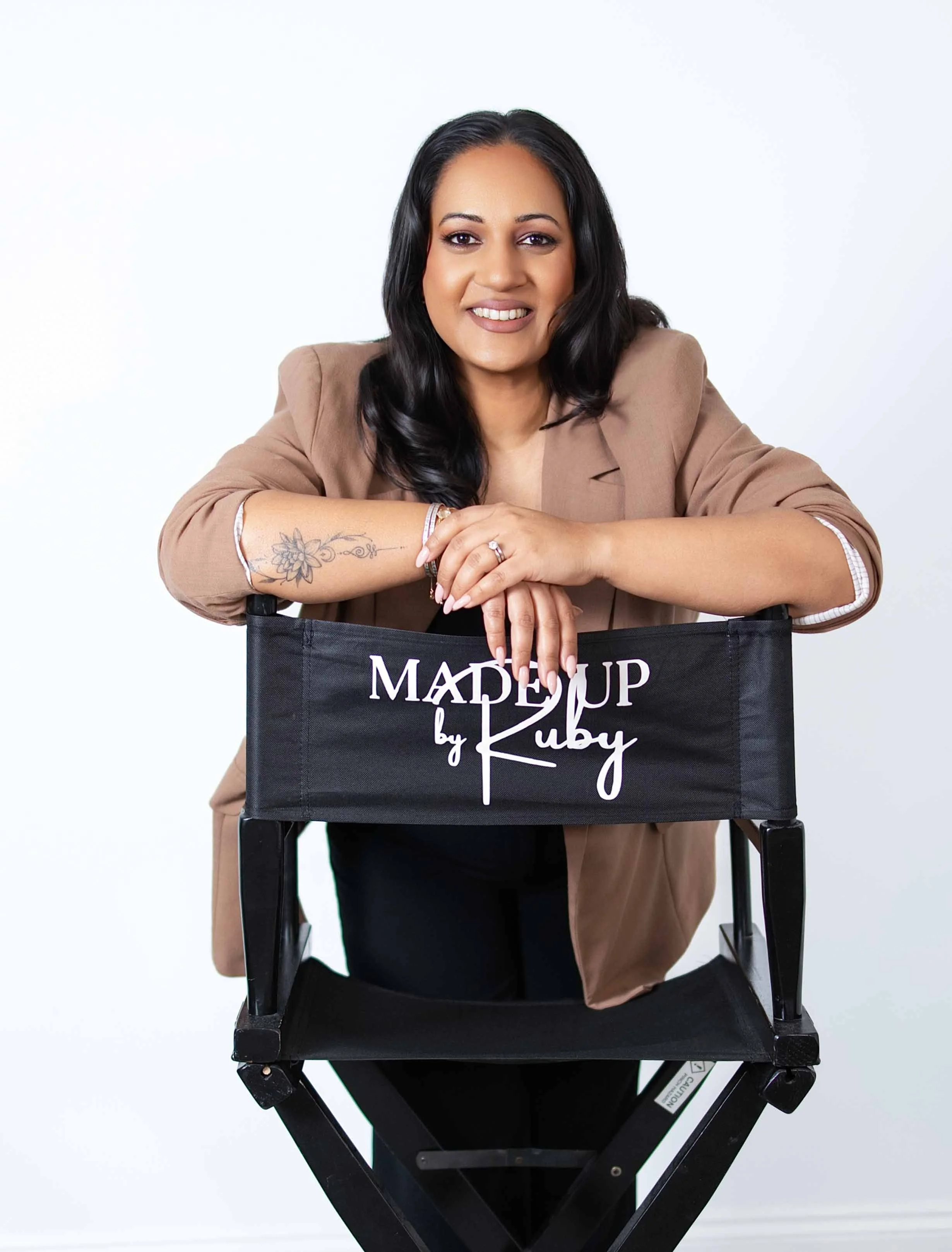A woman with black hair, wearing a tan blazer, leaning on a black director's chair that has a logo reading 'Made Up by Ruby,' smiling at the camera.