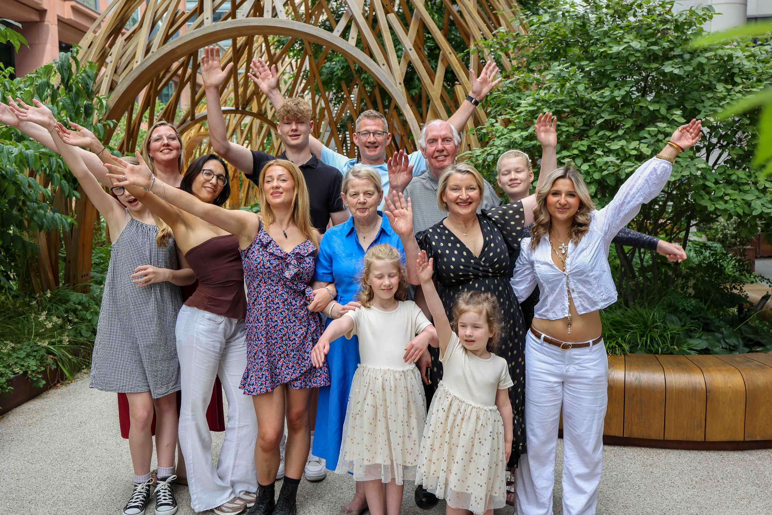 A large, diverse family group posing outdoors in a garden with trees and a wooden arch, smiling and raising their arms.