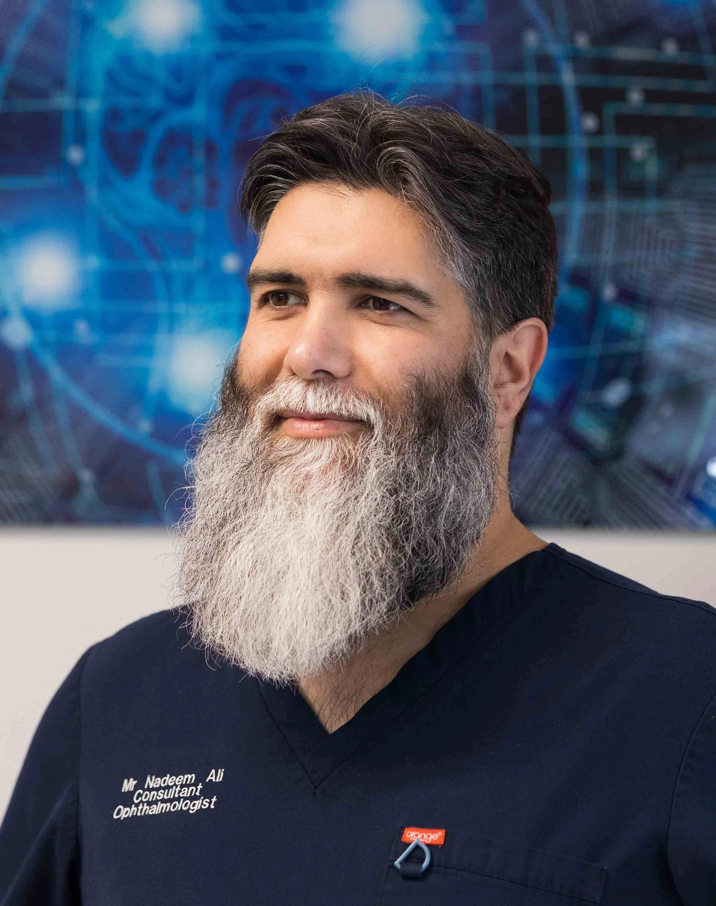 A man with dark hair and a long gray beard wearing navy blue medical scrubs that have his name and title embroidered on them, standing in front of a blue digital background.