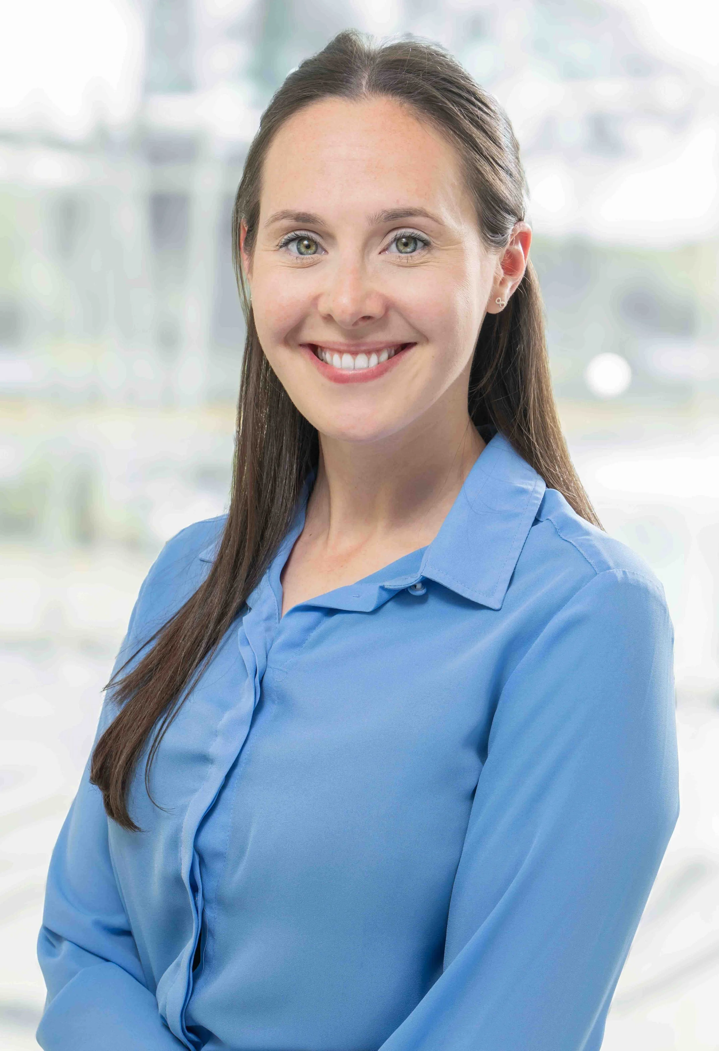 A young woman with long brown hair, wearing a light blue collared shirt, smiling against a blurred background of an office or indoor space.