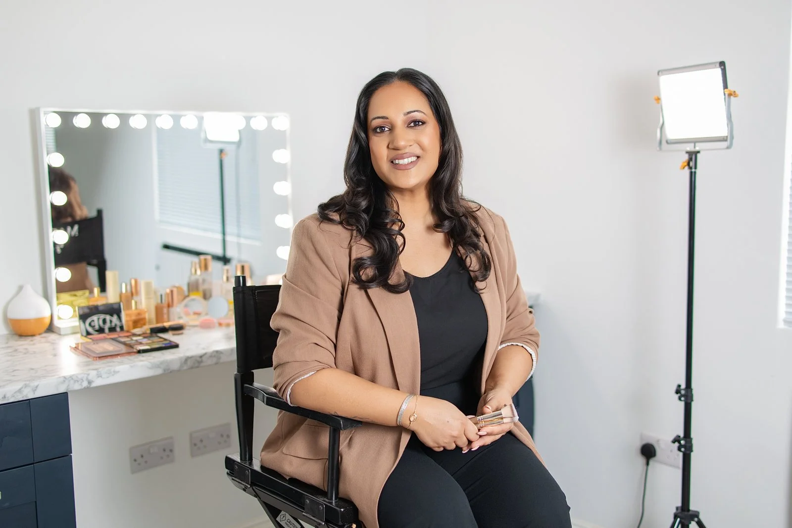 A woman sitting in a makeup chair in front of a mirror with lights, holding a lipstick, in a well-lit room with makeup products on a marble countertop, and professional lighting equipment.